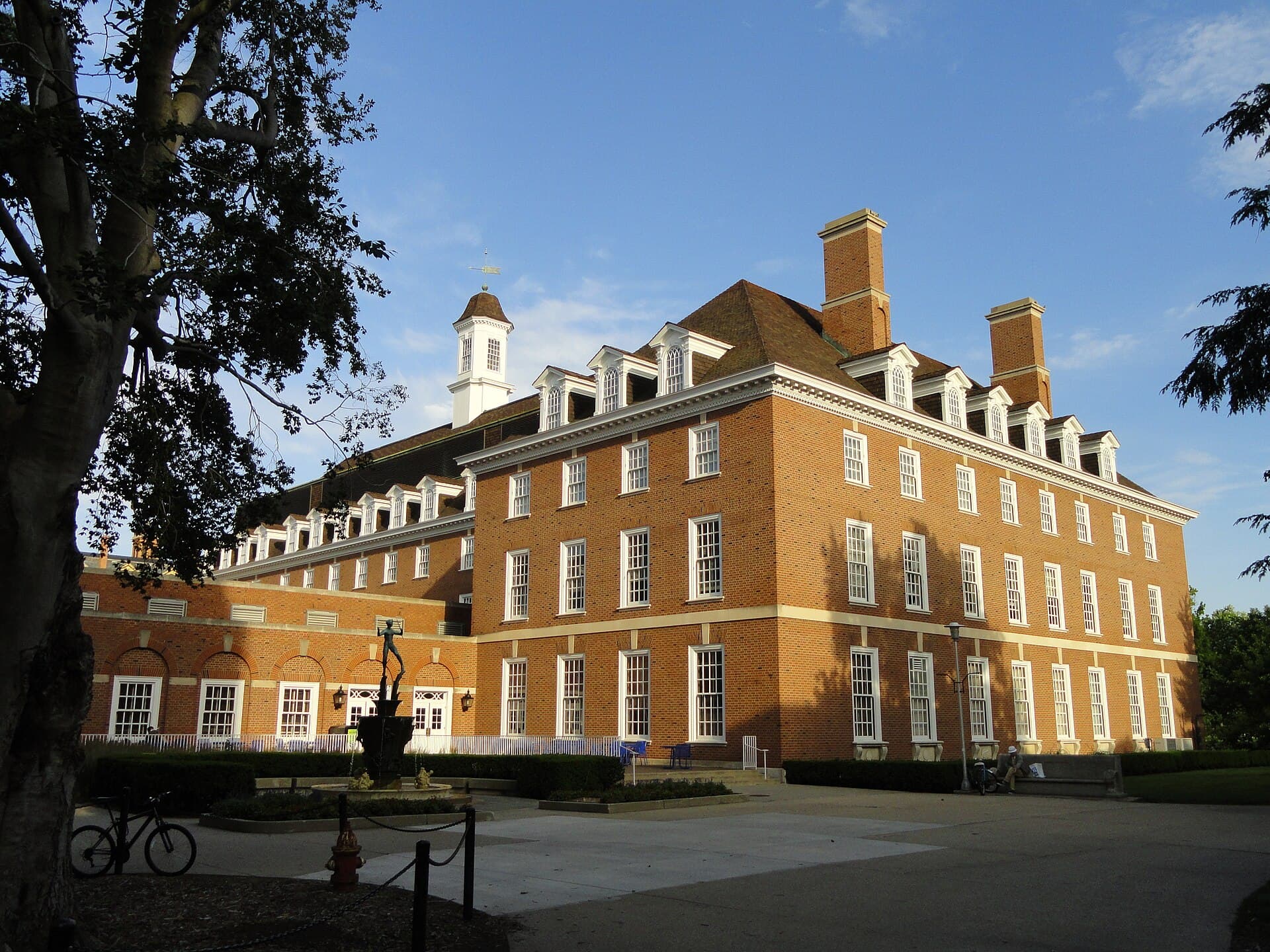 The Illini Union at the University of Illinois, a large red-brick Georgian building with white trim, dormer windows and a white cupola, seen past a tree in evening sun