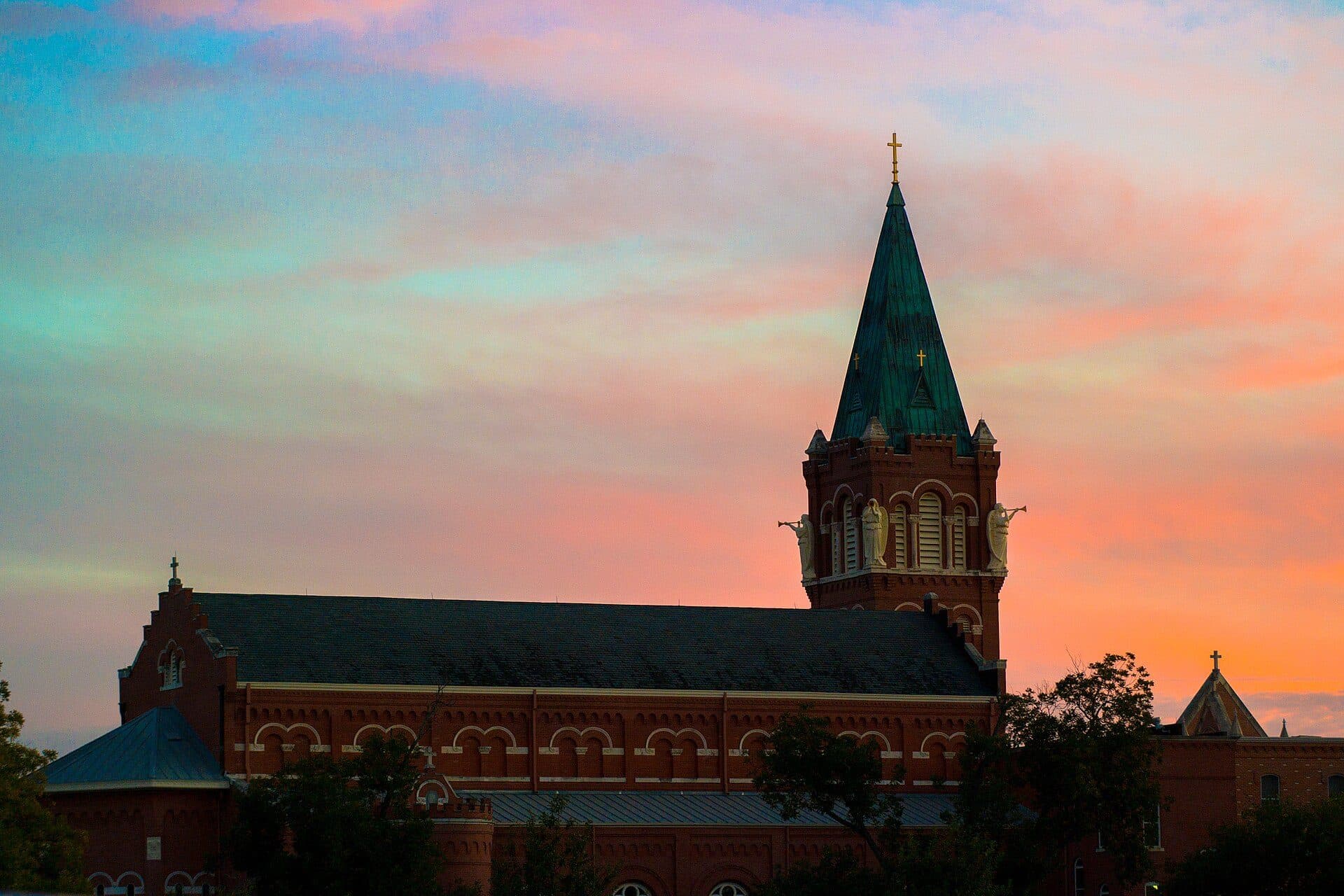 A red-brick building at the University of the Incarnate Word with a tall green spire topped by a cross, silhouetted against a pink and orange sunset sky