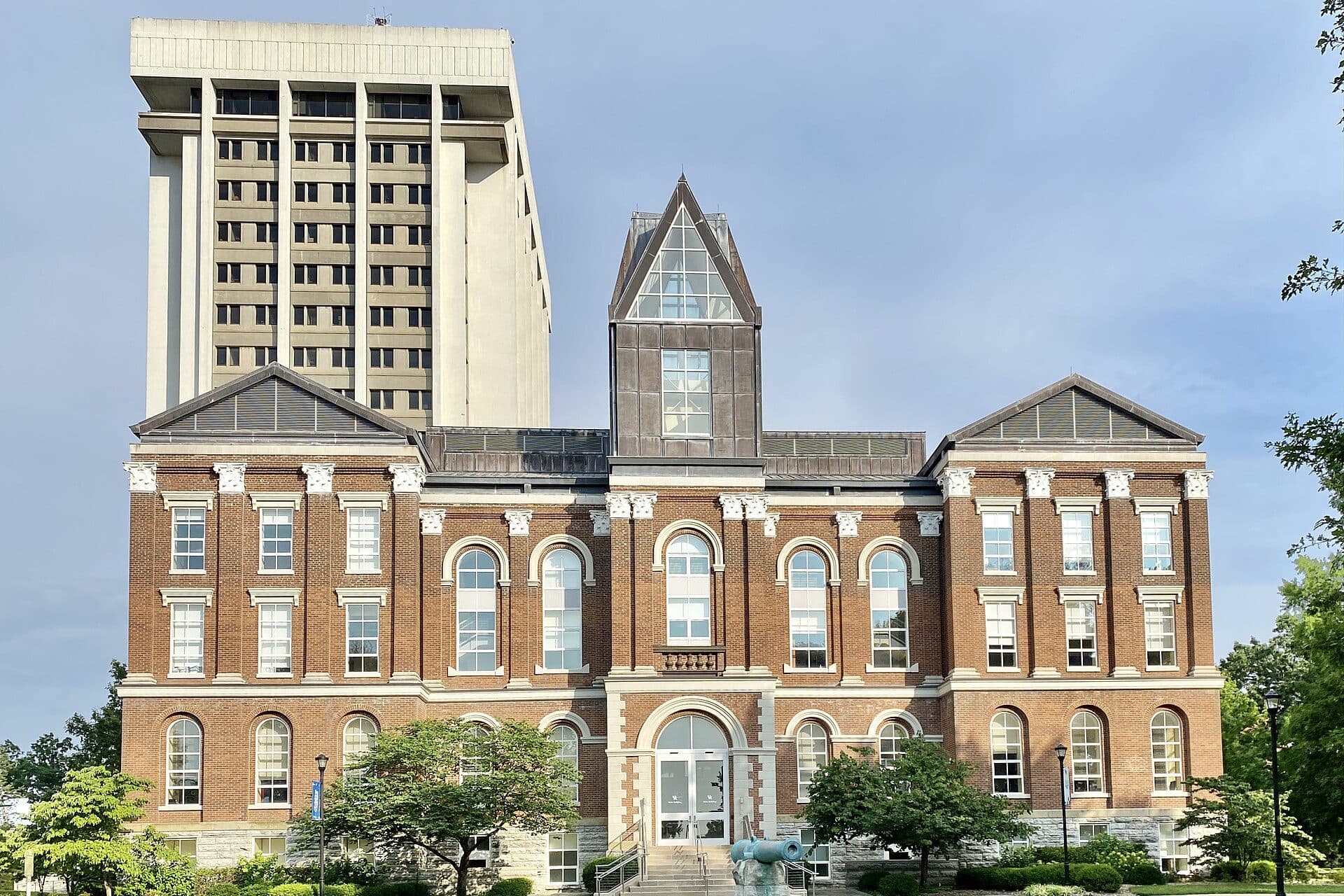 The red-brick, Classical Revival Old Main building with its central gabled tower and white trim, framed by trees and shrubs, with a taller modern building behind it