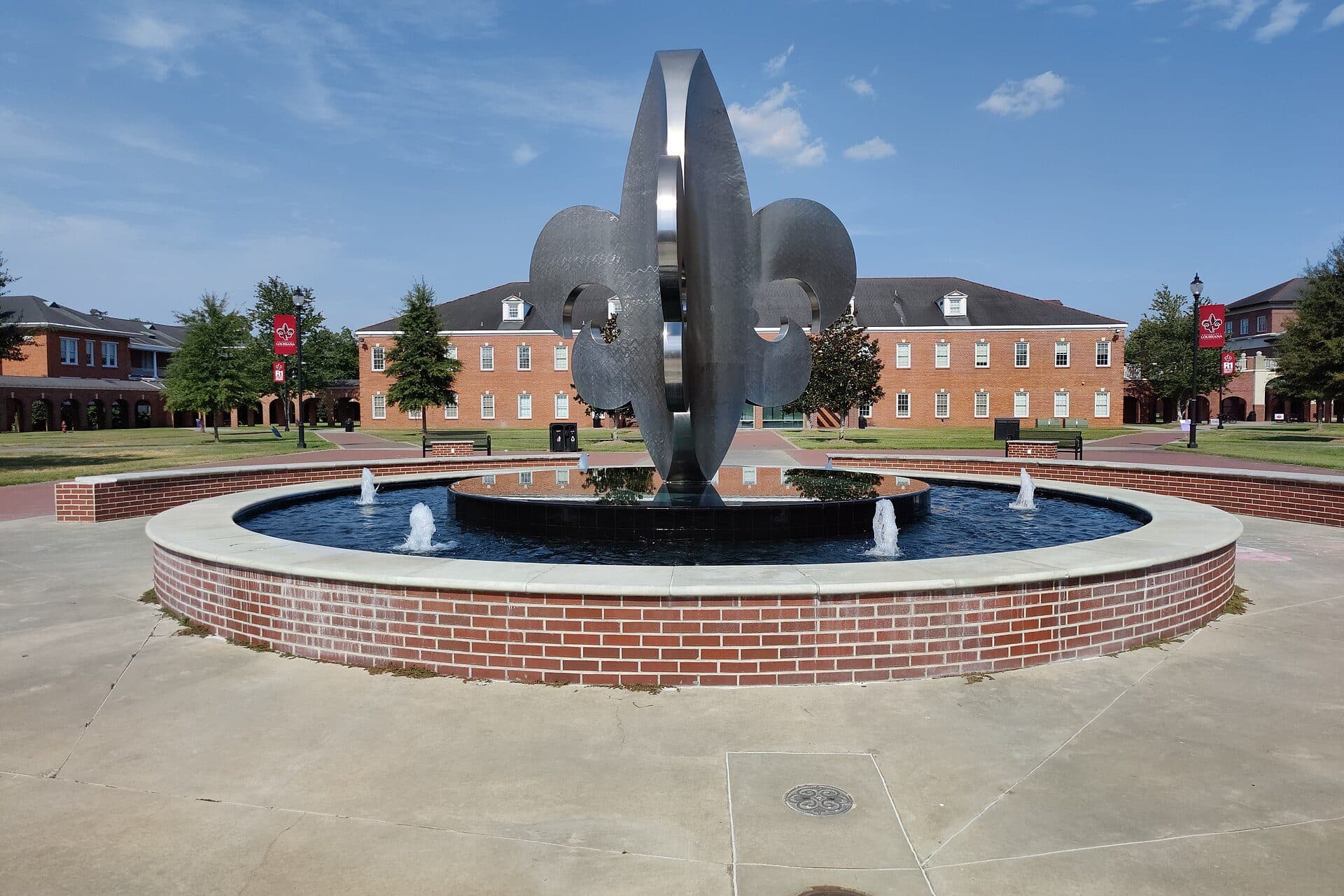 A tall silver fleur-de-lis sculpture rising from a round red-brick fountain in UL Lafayette's Quad, with red-brick buildings and lawns behind it