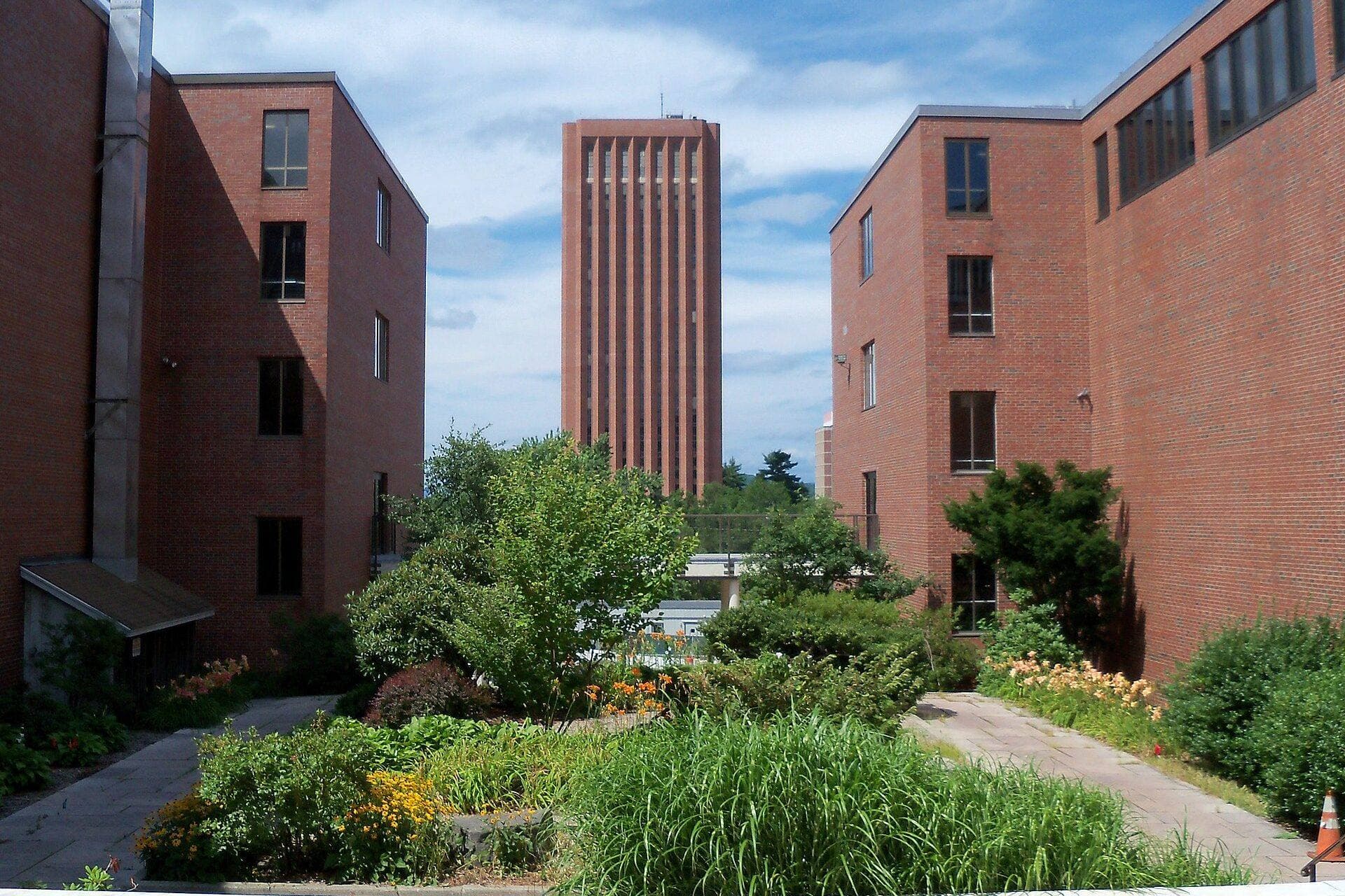 Red-brick UMass Amherst buildings framing a garden path, with the tall brick W.E.B. Du Bois Library tower in the distance under a partly cloudy sky
