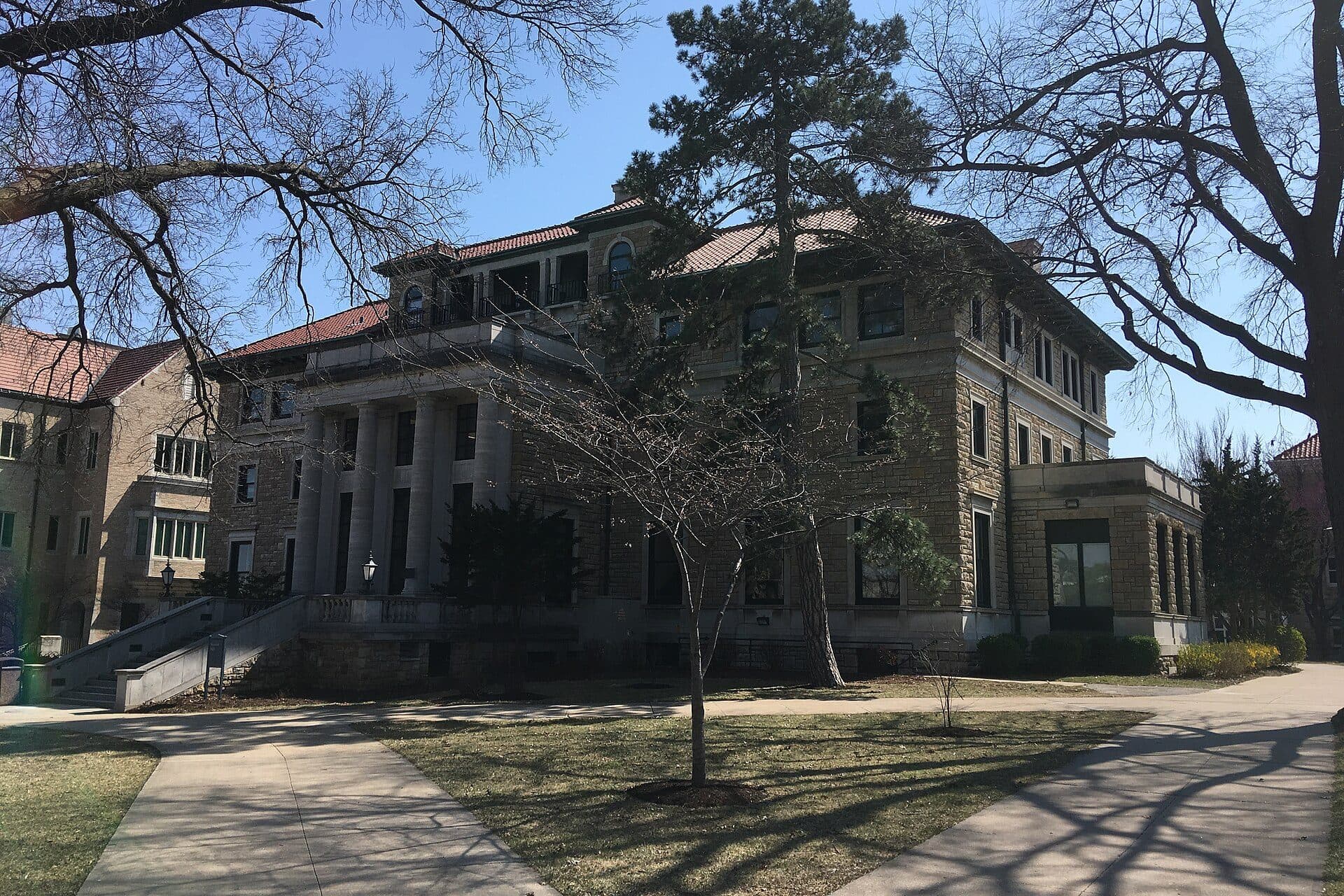 Scofield Hall, a stone building with columns and a red tile roof, seen past bare trees and curving walkways on the UMKC campus