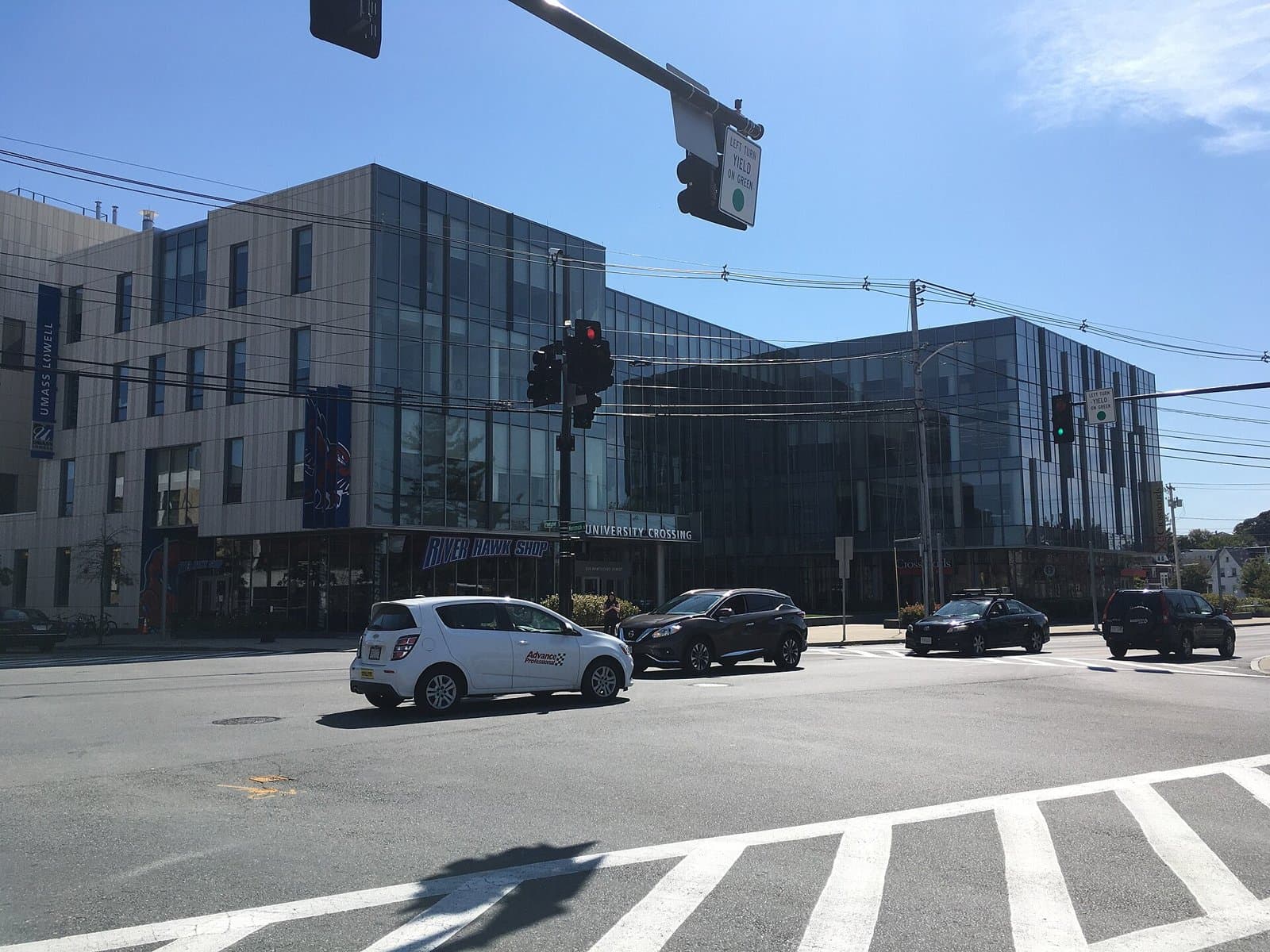 The glass and stone University Crossing building at UMass Lowell, with UMass Lowell banners and the River Hawk Shop, seen across a Lowell intersection on a sunny day