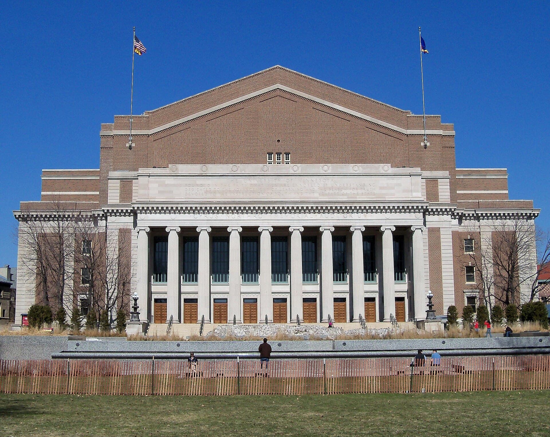 Northrop's brick and limestone facade with tall white columns and two flags under a clear blue sky, seen across the lawn of Northrop Mall