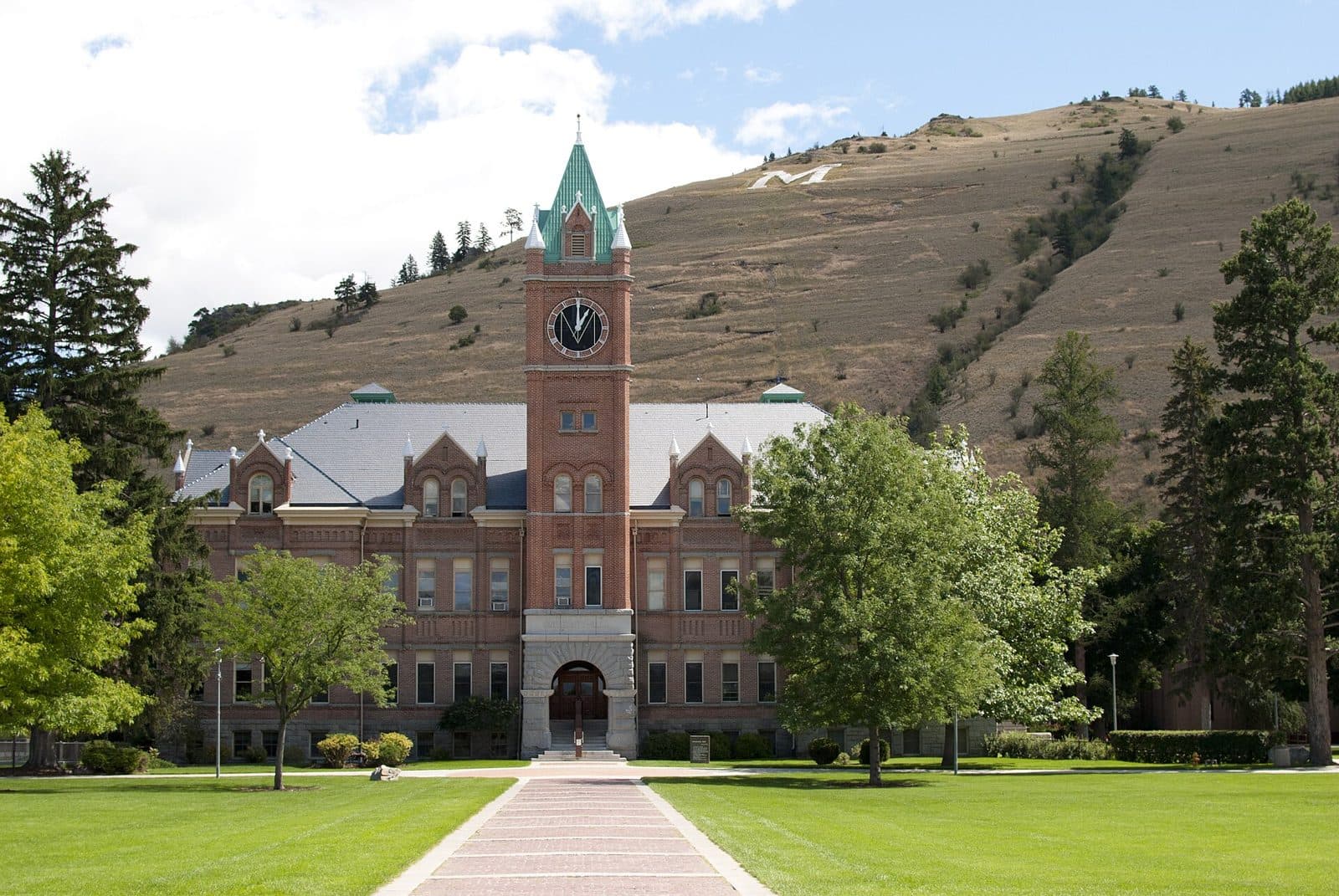 A red-brick hall with a central clock tower topped by a green spire, at the end of a brick walkway across a green lawn, with a grassy mountainside marked by a large white M rising behind it under a partly cloudy sky