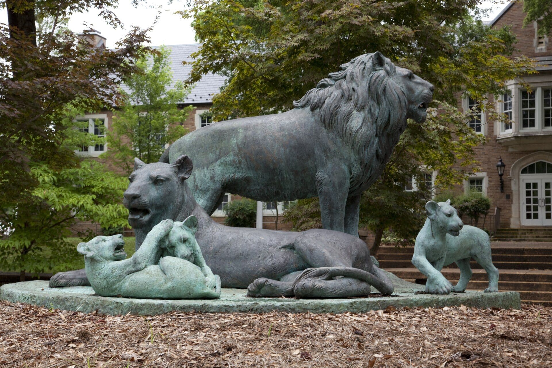 A bronze sculpture of a standing lion, a lioness and three cubs on a round base in front of a red-brick building with white-trimmed windows, framed by green trees