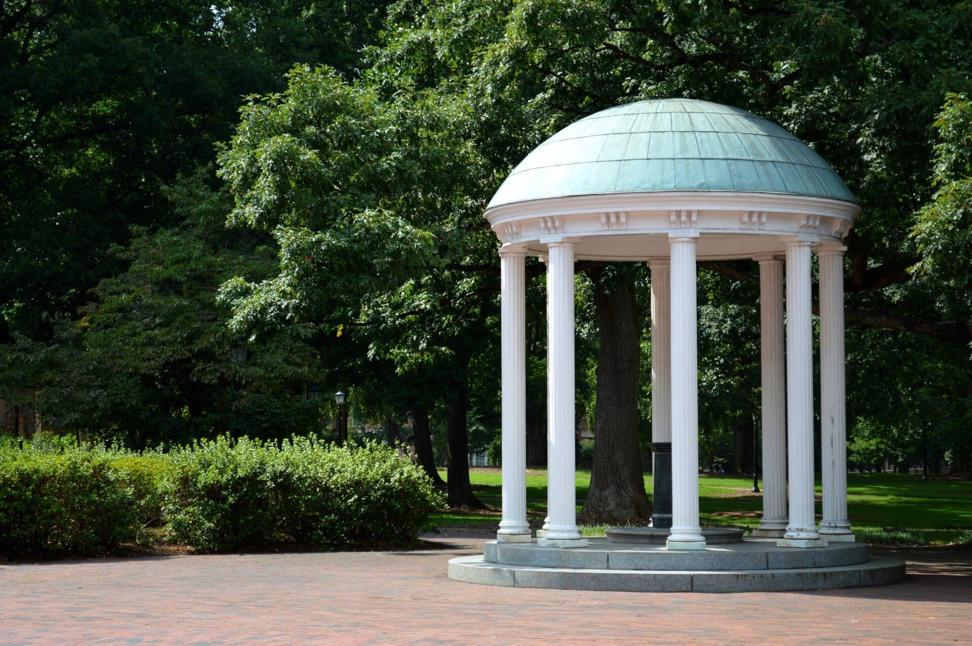 The Old Well at UNC Chapel Hill, a small white-columned rotunda with a green dome on a brick plaza, surrounded by tall shade trees