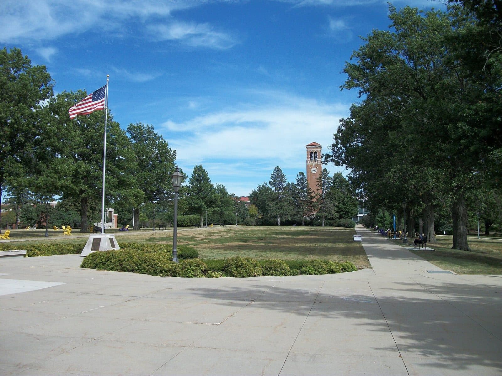 A wide plaza and tree-lined lawn at the University of Northern Iowa leading to the brick Campanile, with an American flag on the left under a blue sky