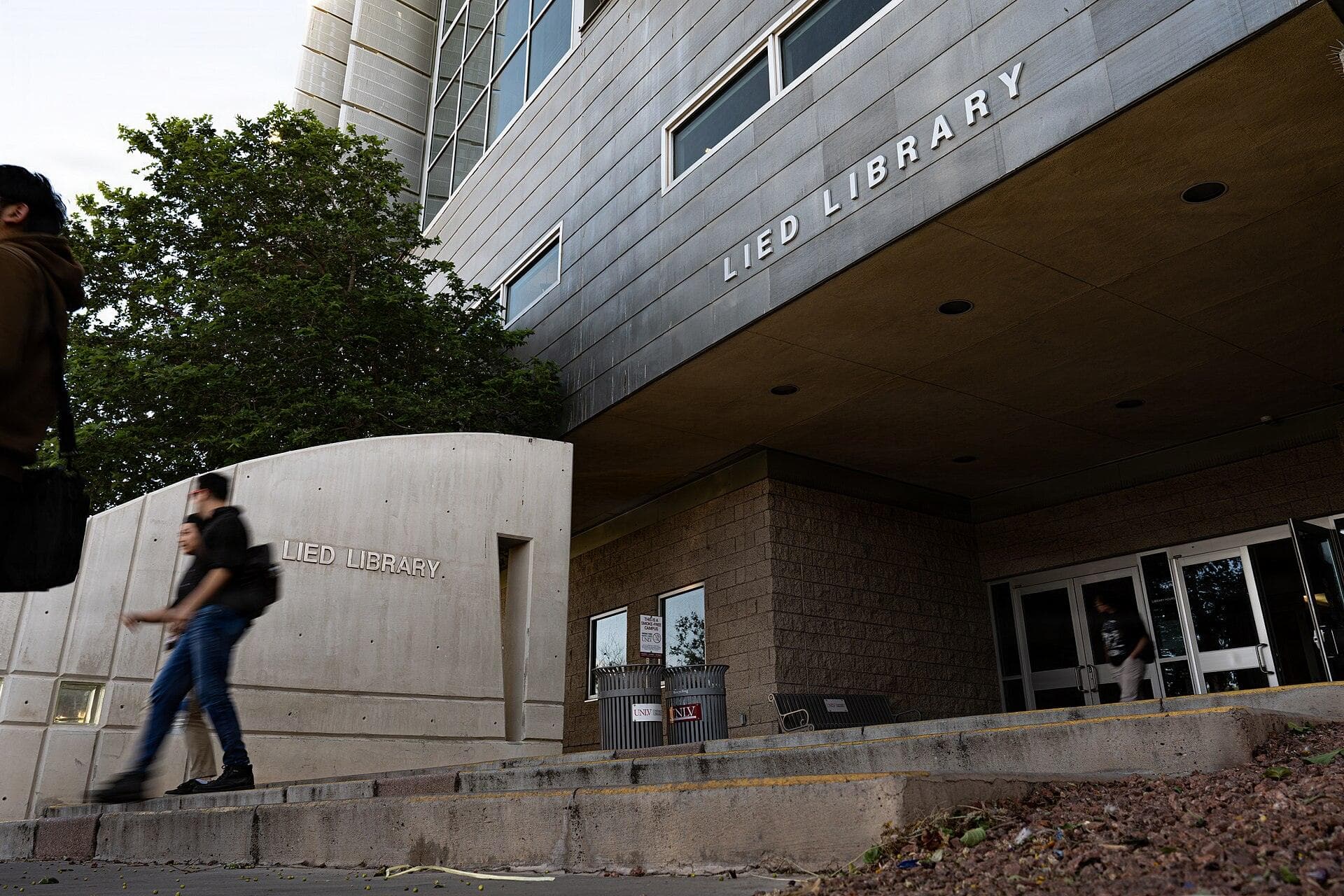 Students walk past the entrance of the Lied Library at UNLV, a modern building with gray metal panels and the words Lied Library above a shaded entry, with a curved concrete wall and a leafy tree