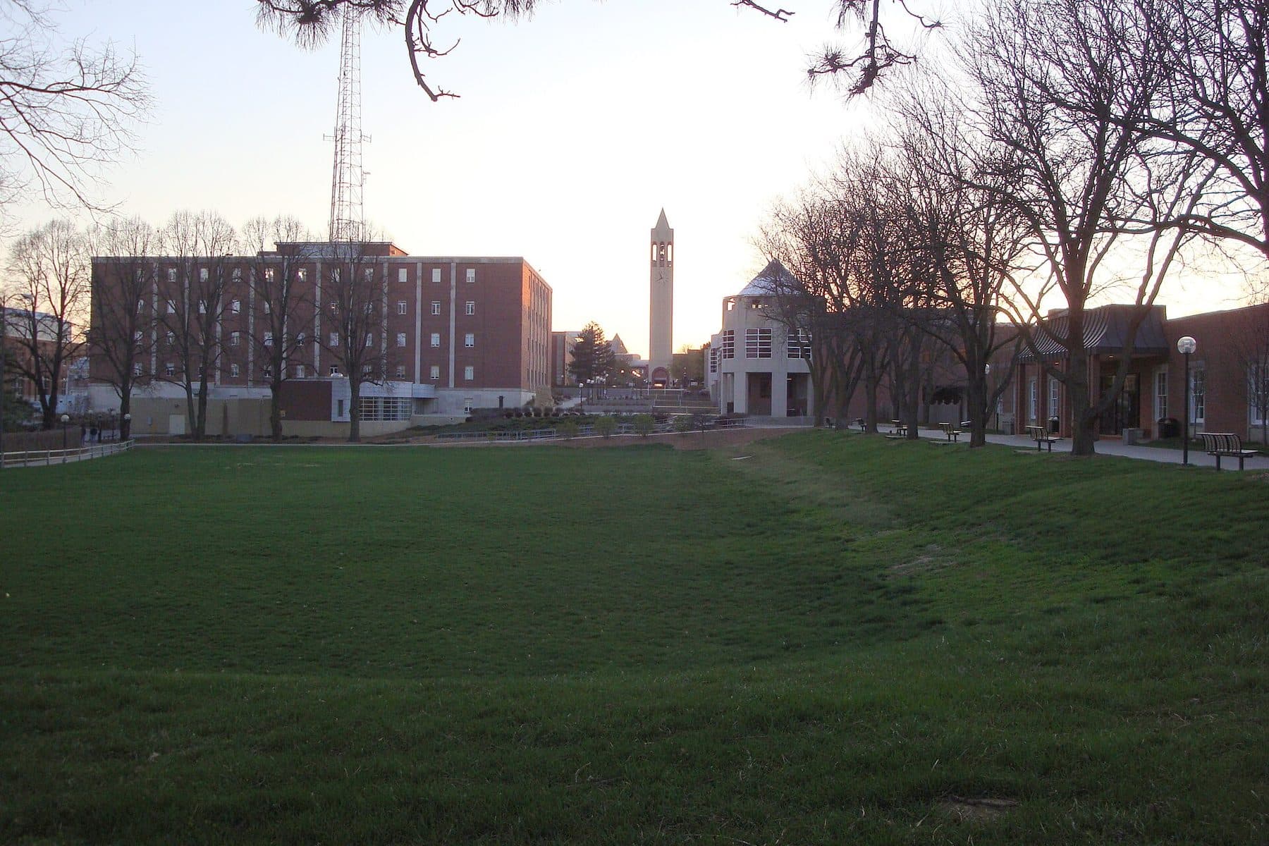 A grassy campus lawn at dusk with brick buildings and a slender tower at the University of Nebraska Omaha