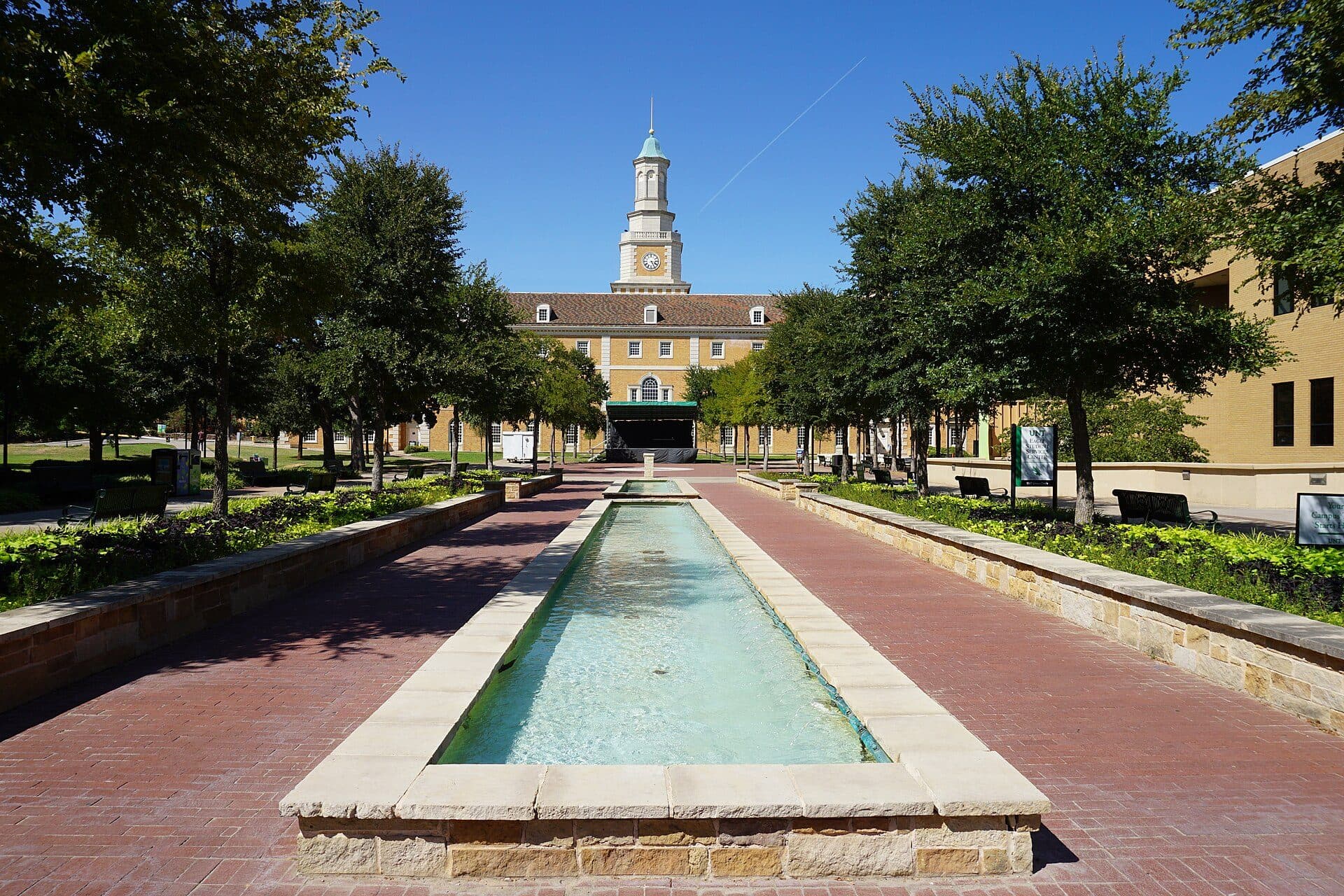 A long reflecting pool lined with brick walks and trees leading to the Hurley Administration Building and its white cupola tower at UNT