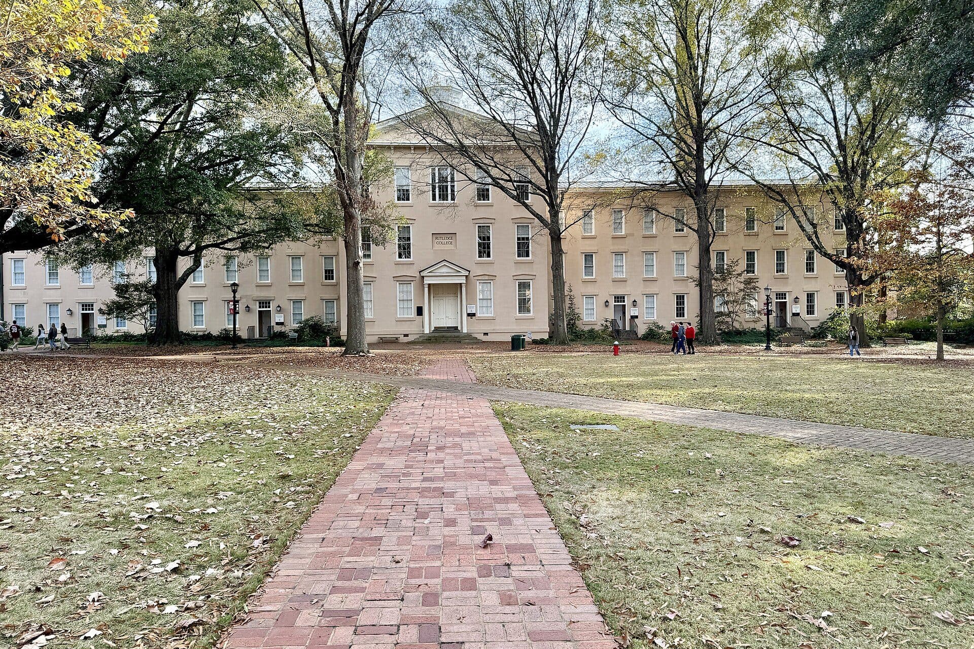 A brick path across the Horseshoe lawn toward Rutledge College, a long cream-colored Federal-style building framed by tall trees at the University of South Carolina