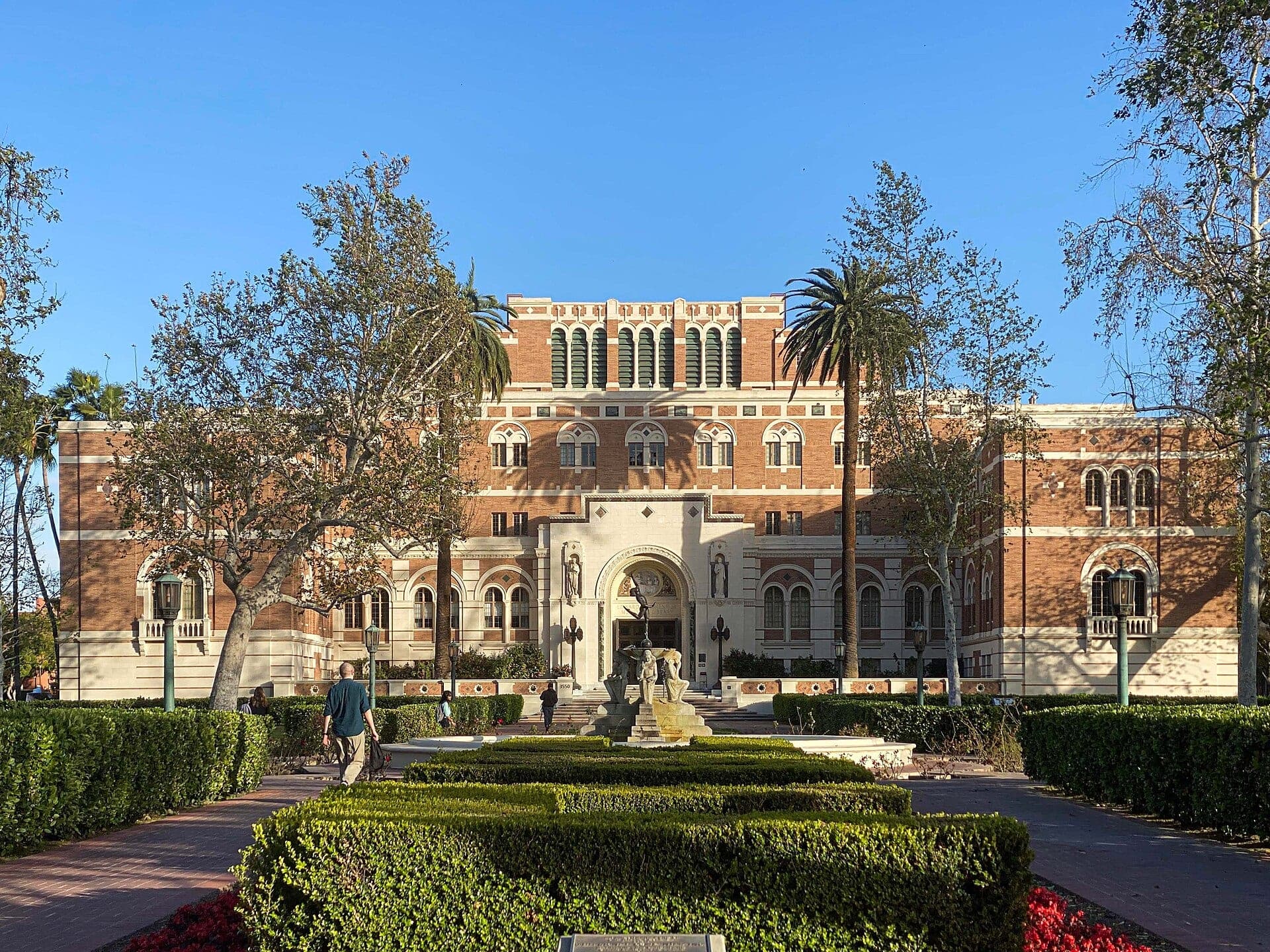 The red-brick Doheny Memorial Library at USC, seen down a hedged walkway lined with trees and a palm on a sunny day