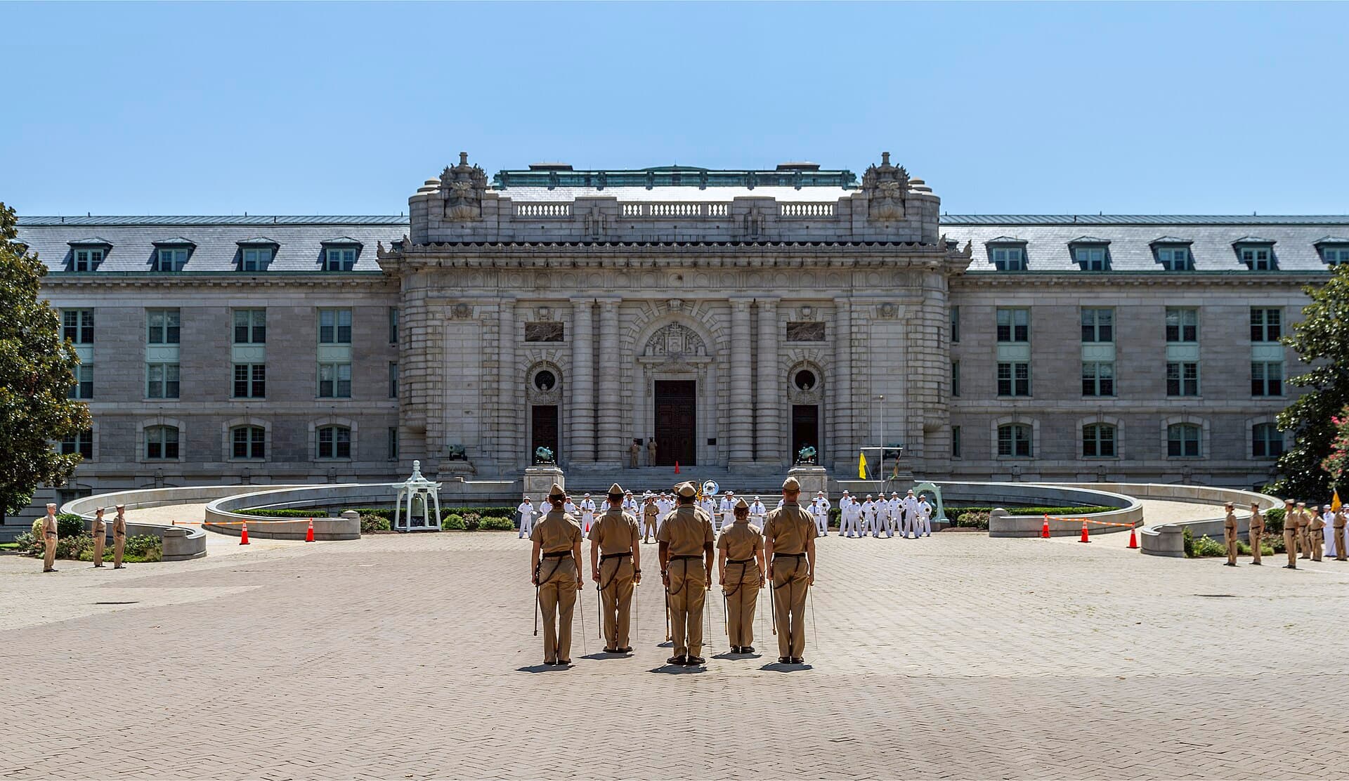 Midshipmen in khaki and white uniforms standing in formation on a brick plaza in front of Bancroft Hall, a long gray granite building with a columned central entrance, at the U.S. Naval Academy