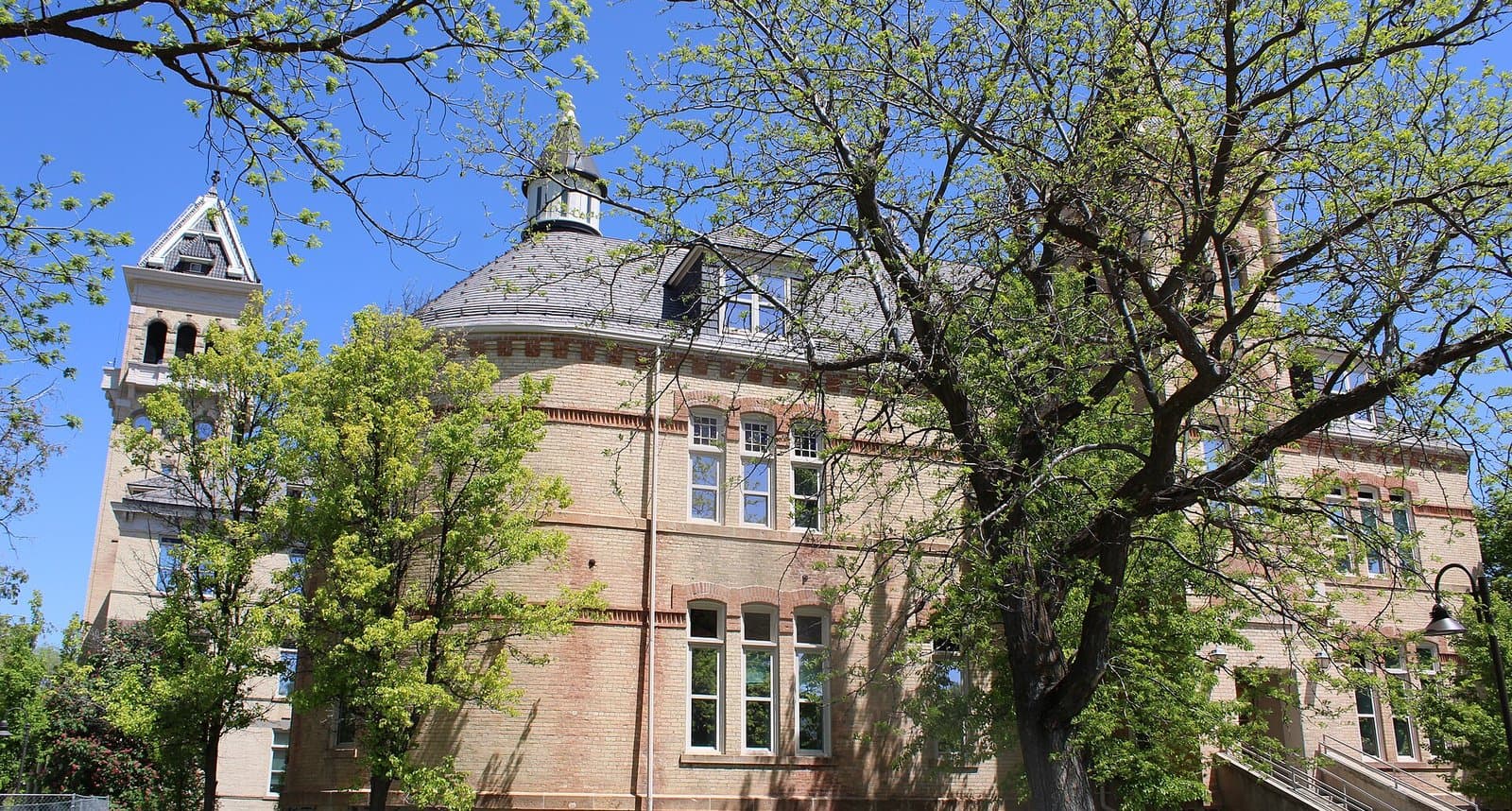 The tan-brick Old Main building with its bell tower and slate roof, framed by leafy spring trees under a clear blue sky at Utah State University