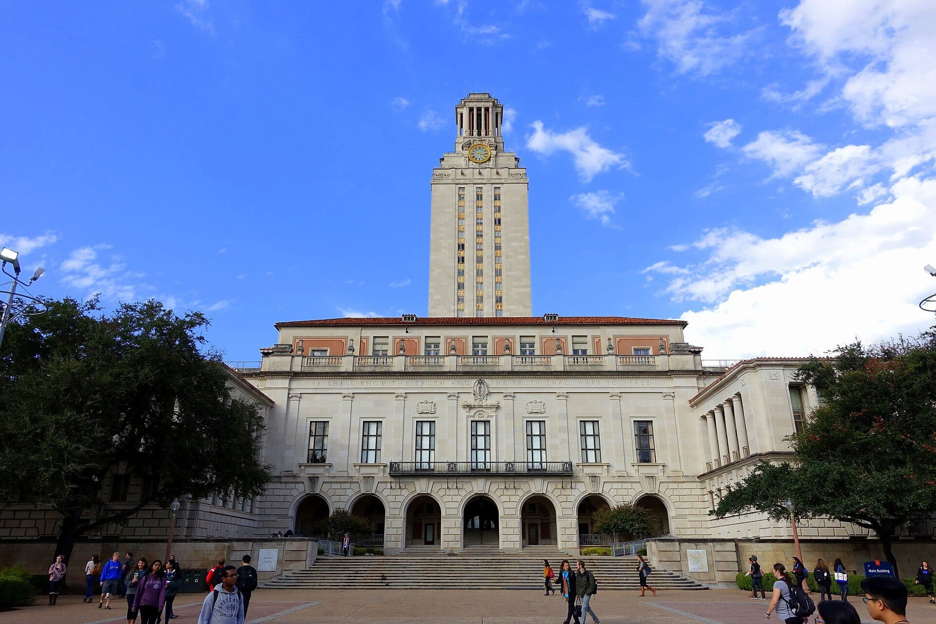 The University of Texas at Austin's Main Building, a pale limestone building with arched doorways and a tall tower rising from its center, seen across a plaza with students walking under a blue sky
