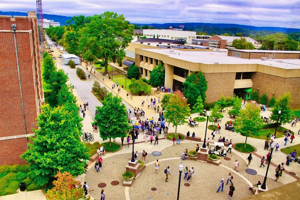 Students crossing a brick plaza and tree-lined walkways at UTC, seen from above, with campus buildings and green ridges behind