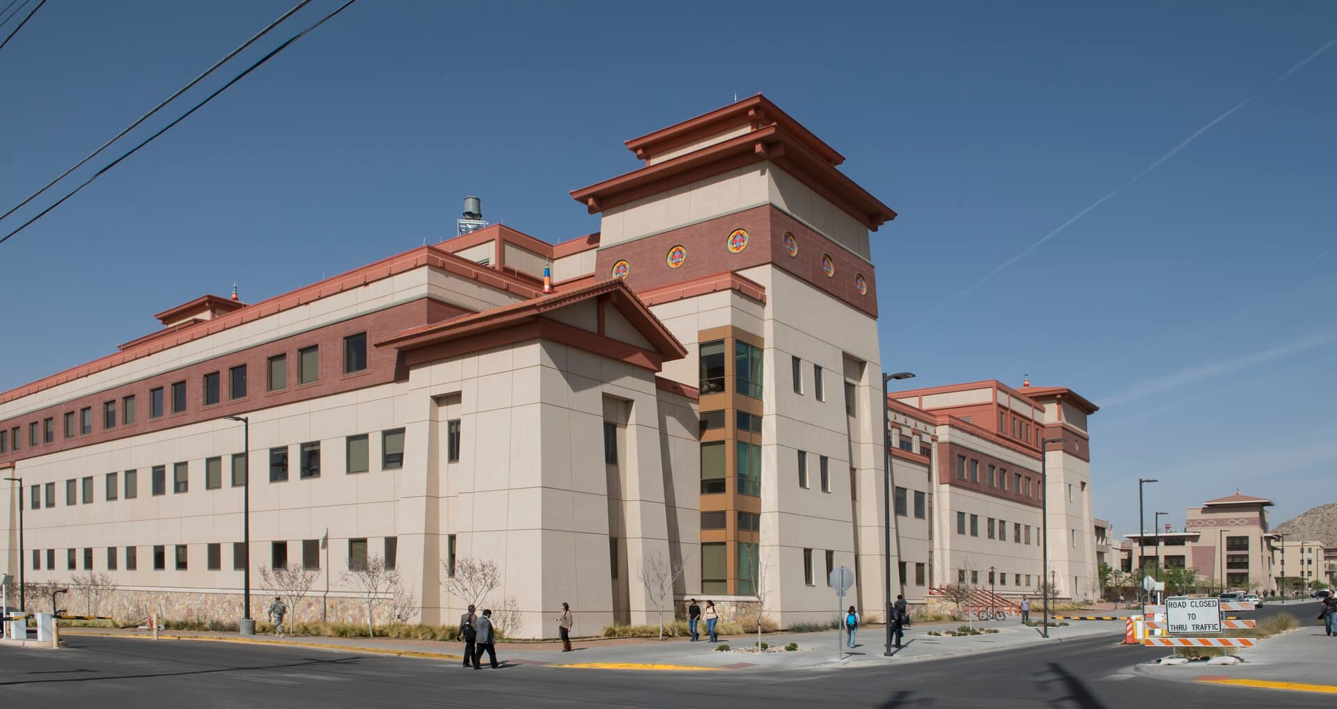 A long Bhutanese-style UTEP building with a cream facade, a dark red band with round medallions and a flared red roof on its corner tower, with students walking on the sidewalk under a clear blue sky