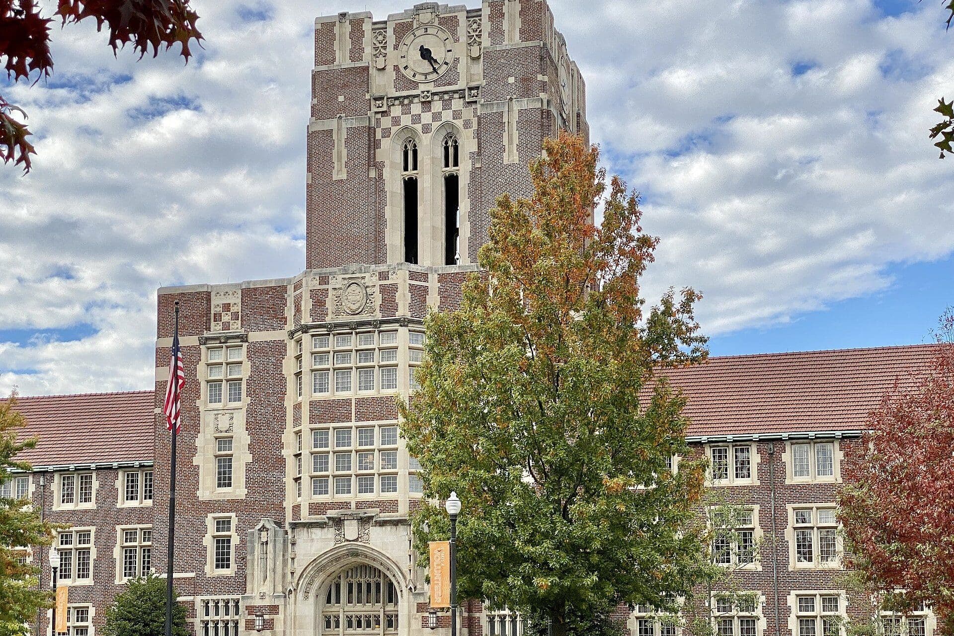 The red-brick Collegiate Gothic clock tower of Ayres Hall rising above autumn trees under a blue sky, with an American flag in front