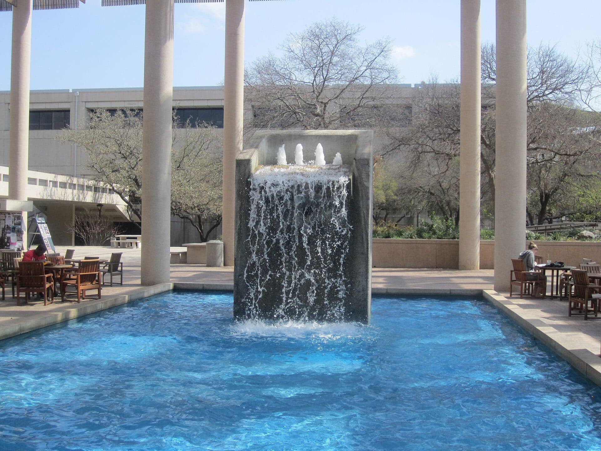Sombrilla Plaza at UTSA's Main Campus: a blue fountain pool with a square waterfall, framed by tall white columns, with tables and chairs and campus buildings behind