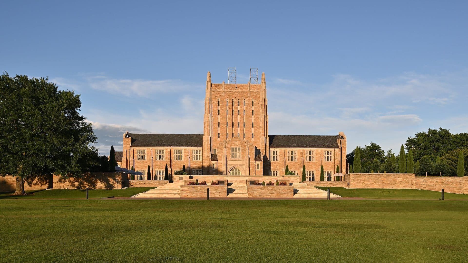 A tall sandstone-colored Gothic-style library with a central tower and long wings, seen across a wide green lawn with broad stone steps and low walls in front, trees at both sides and a blue sky with light clouds