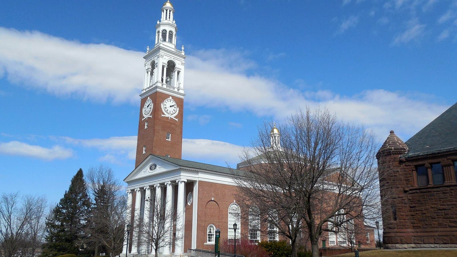 A red-brick chapel with white columns and a tall brick bell tower topped by a white clock-and-cupola spire, beside bare trees and a stone building under a blue sky with white clouds