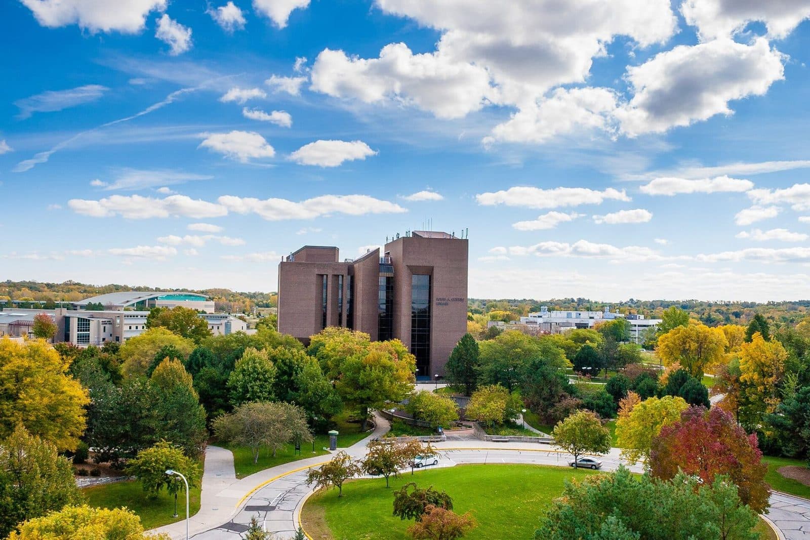 A tall brown-brick library tower rising behind trees in orange and yellow fall color, with a curving campus drive and green lawn in front under a blue sky with puffy white clouds