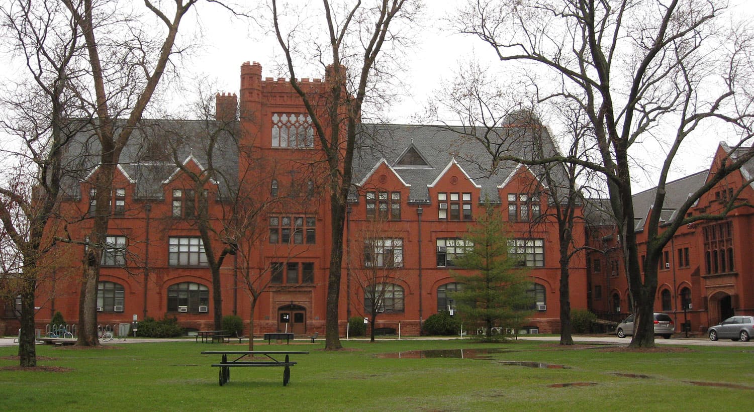 A long red-brick Gothic-style hall with a crenellated tower and gray slate gables at UW-Milwaukee, seen across a green lawn with picnic tables and tall bare trees on an overcast spring day