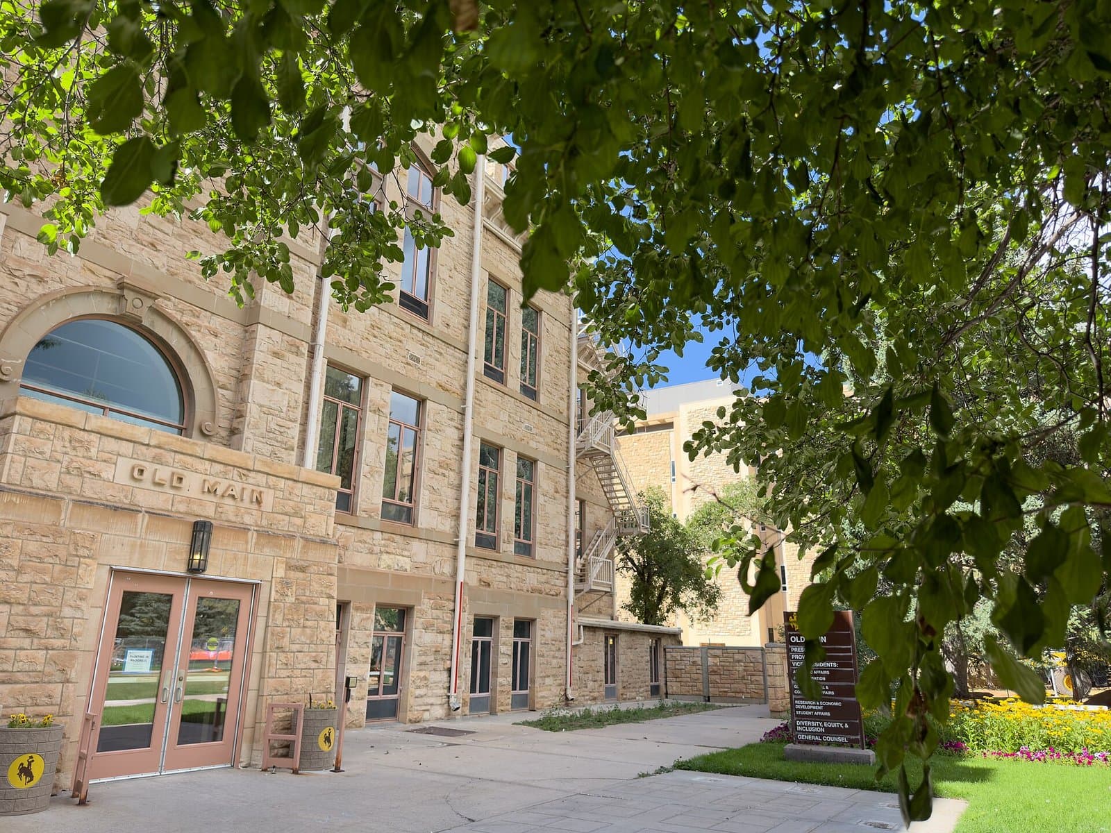 The pale sandstone Old Main building with OLD MAIN carved over a pair of glass doors, a large arched window and rows of tall windows, seen in summer under the leafy branches of a tree, with flower beds and a lawn to the right under a blue sky