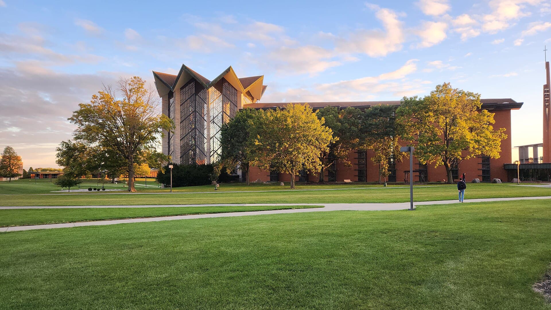 A tall chapel with a peaked, glass-walled front and a long red-brick wing behind a row of autumn trees, seen across a wide green lawn and curving sidewalk under a blue sky with pink-tinted clouds