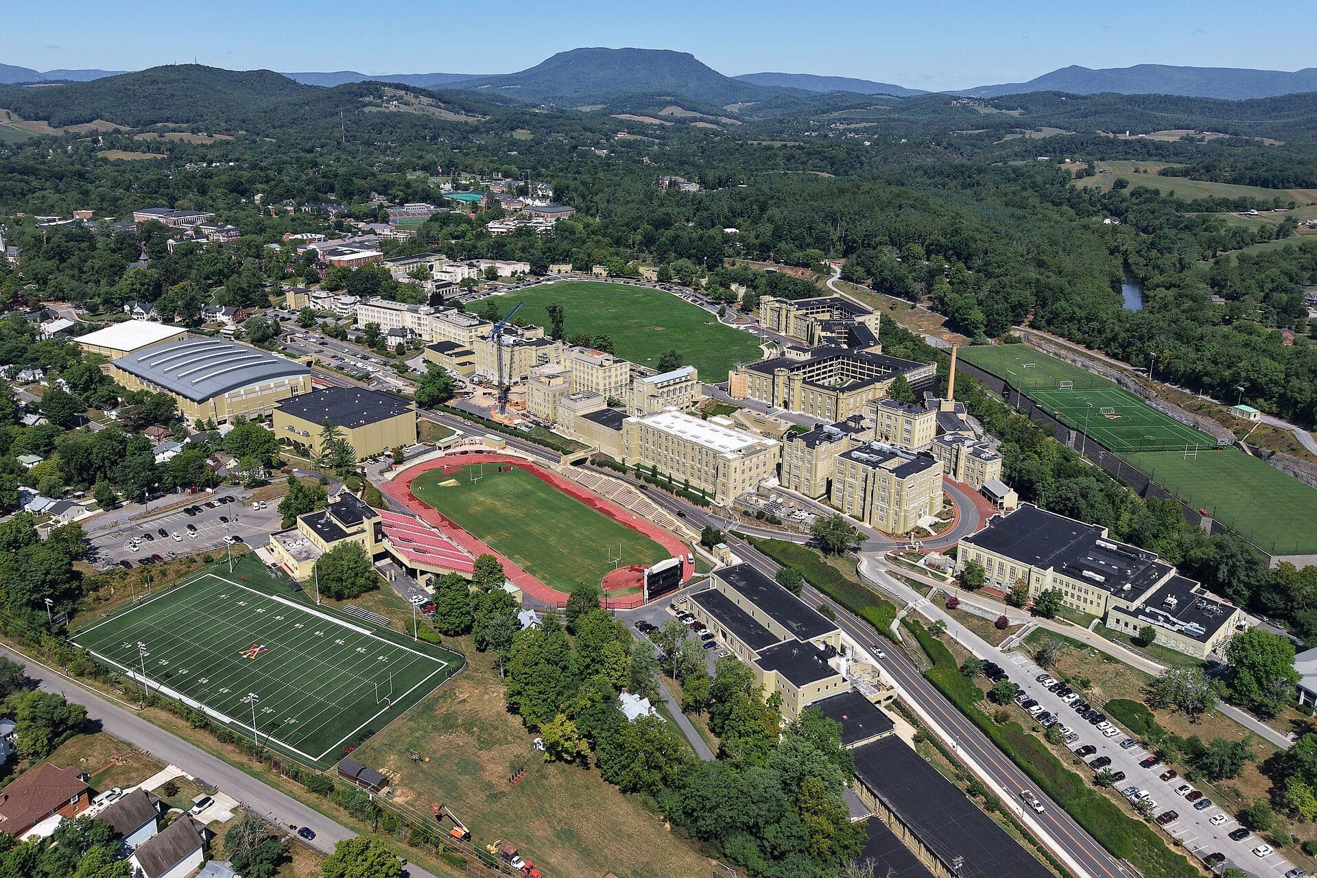 Aerial view of the VMI post in Lexington, with pale barracks and academic buildings, a parade ground, a running track and athletic fields, and wooded mountains behind