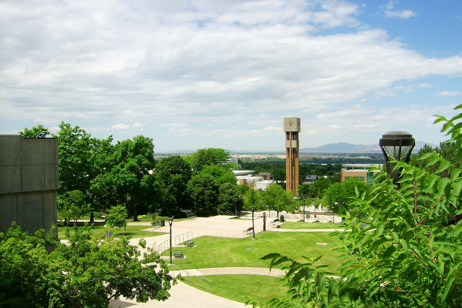 A green, tree-lined Weber State campus walkway leading toward the bell tower, with the valley beyond