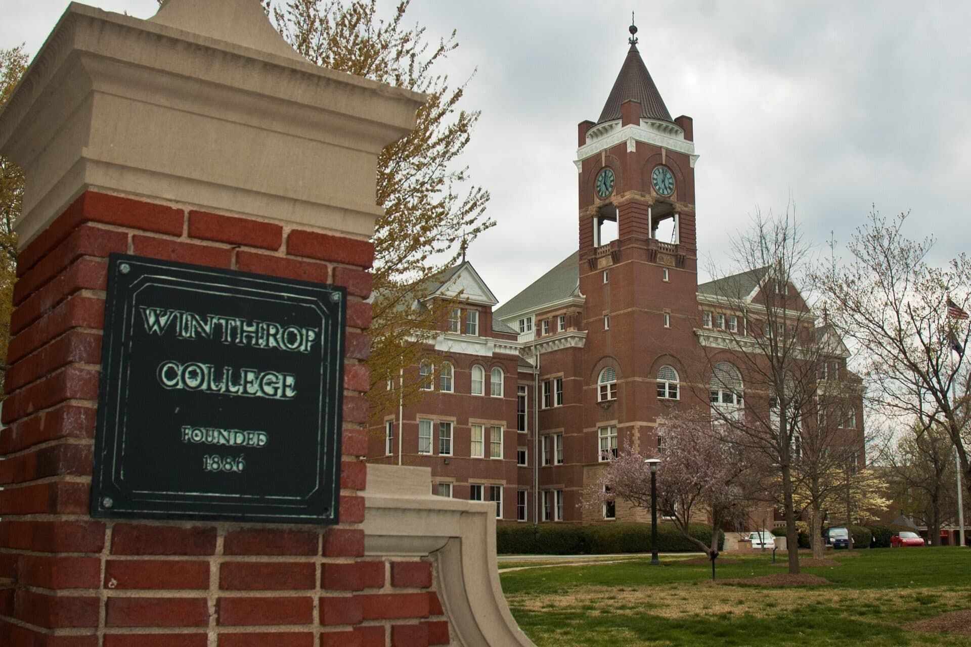 A brick gate pillar with a Winthrop College plaque in front of Tillman Hall's red-brick clock tower at Winthrop University
