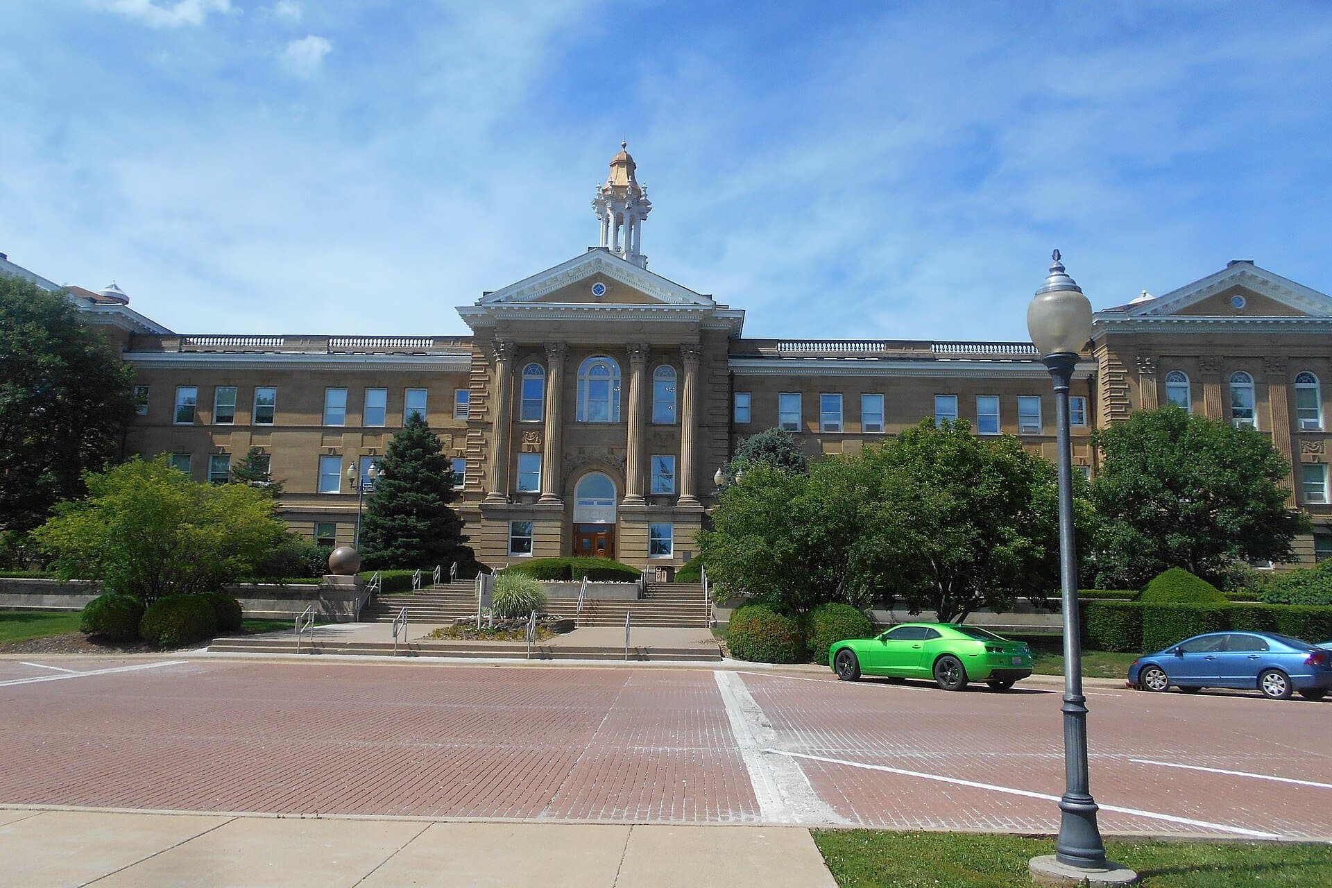 A large tan brick building with columns and a white cupola, behind a brick plaza with a lamppost and parked cars under a blue sky