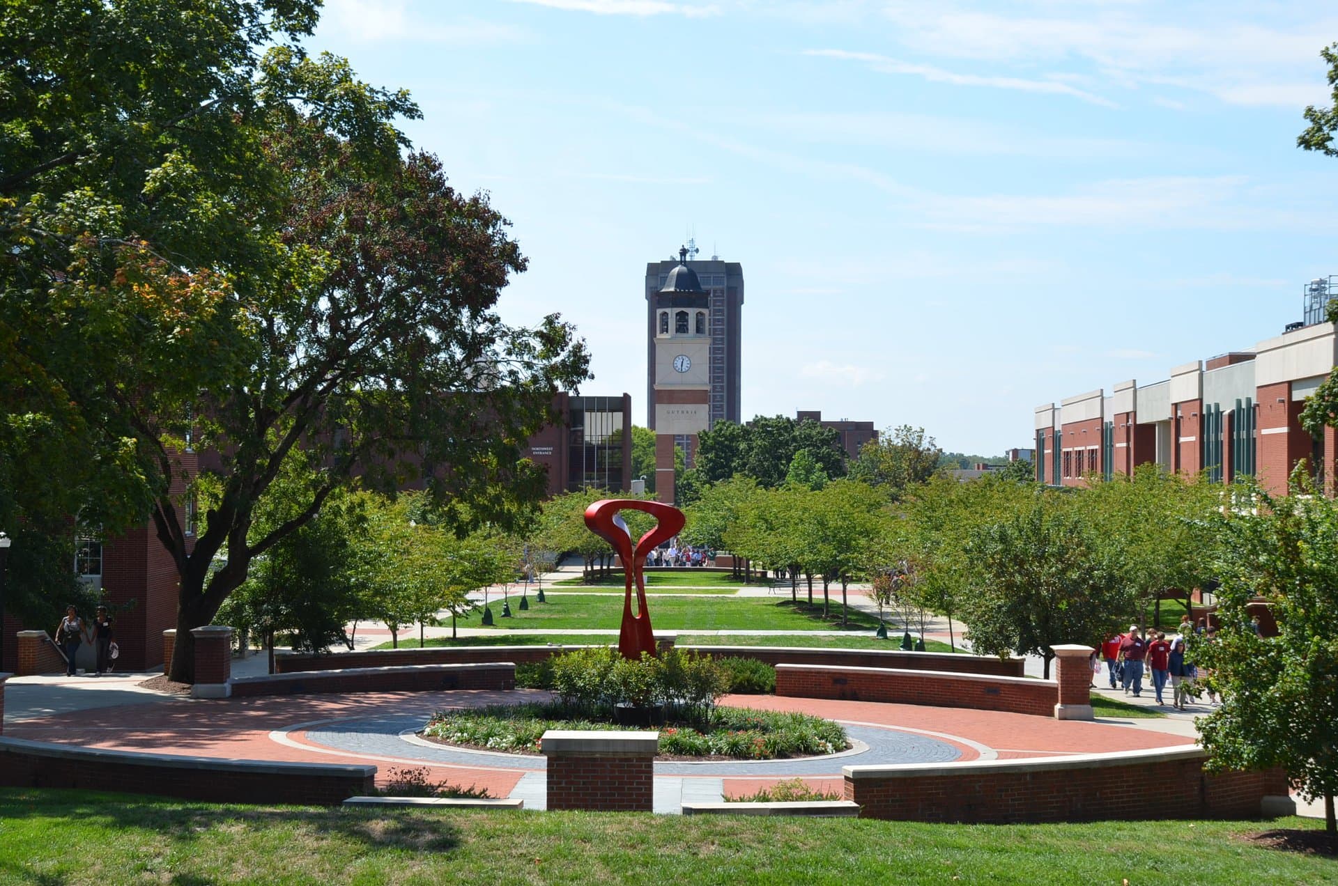 A red curving sculpture in a round brick plaza at the center of Western Kentucky University's campus, with a long green lawn, young trees and a brick clock tower in the distance under a blue sky