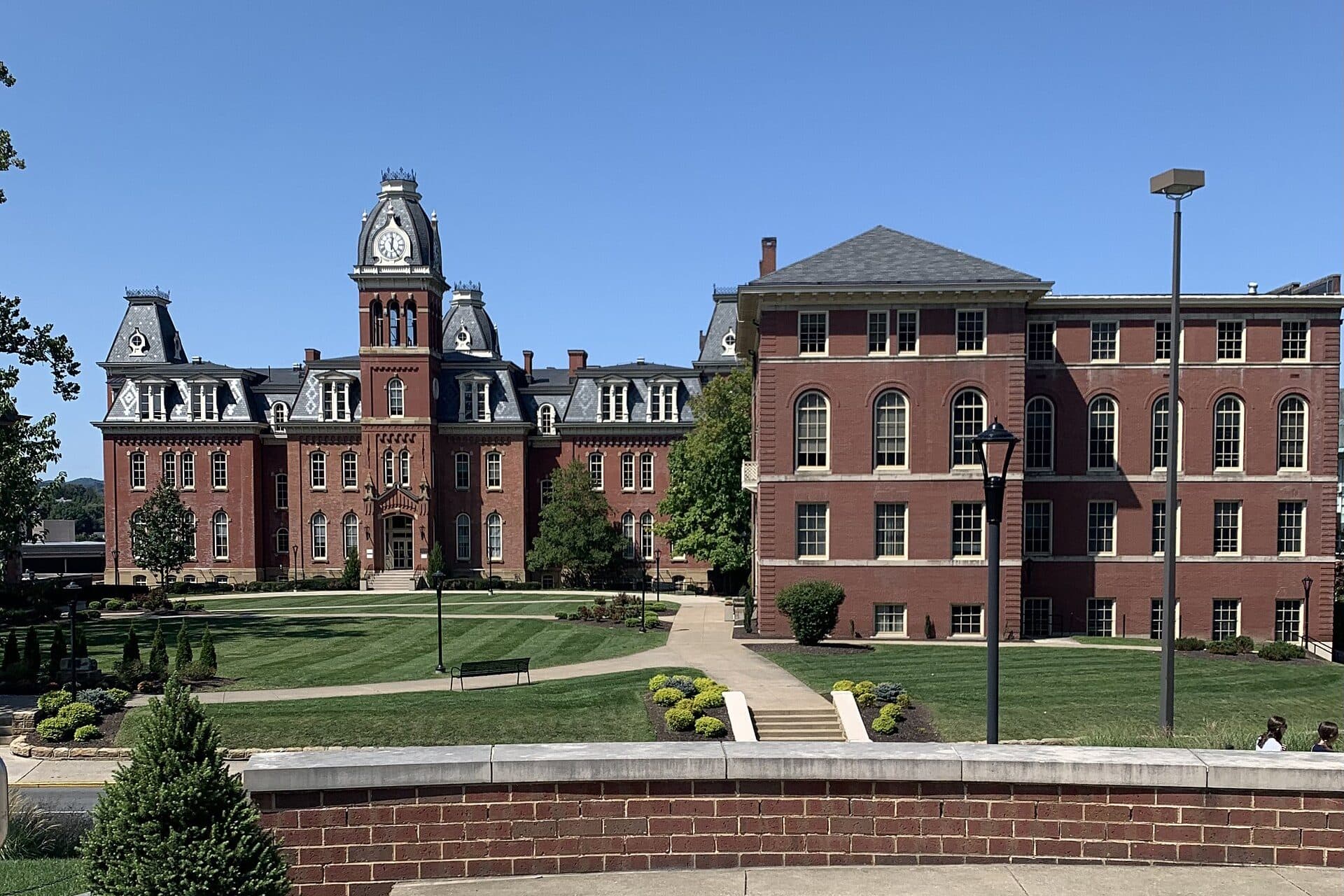 Red-brick Woodburn Hall with its central clock tower and mansard roof behind a landscaped lawn and brick wall on WVU's Downtown Campus