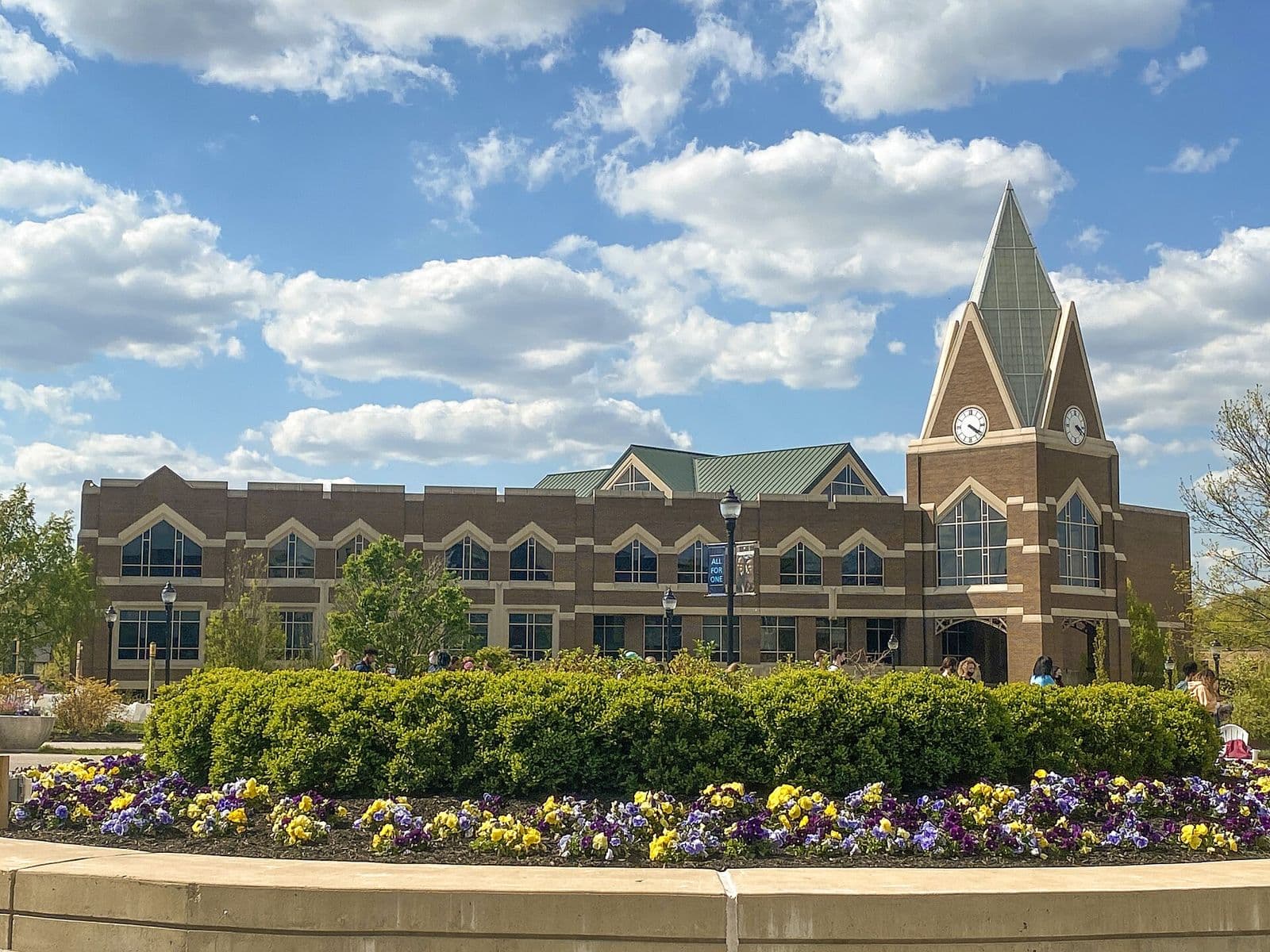 A brick student center with arched windows and a clock tower topped by a green spire, behind a hedge and a bed of yellow and purple pansies under a blue sky with white clouds