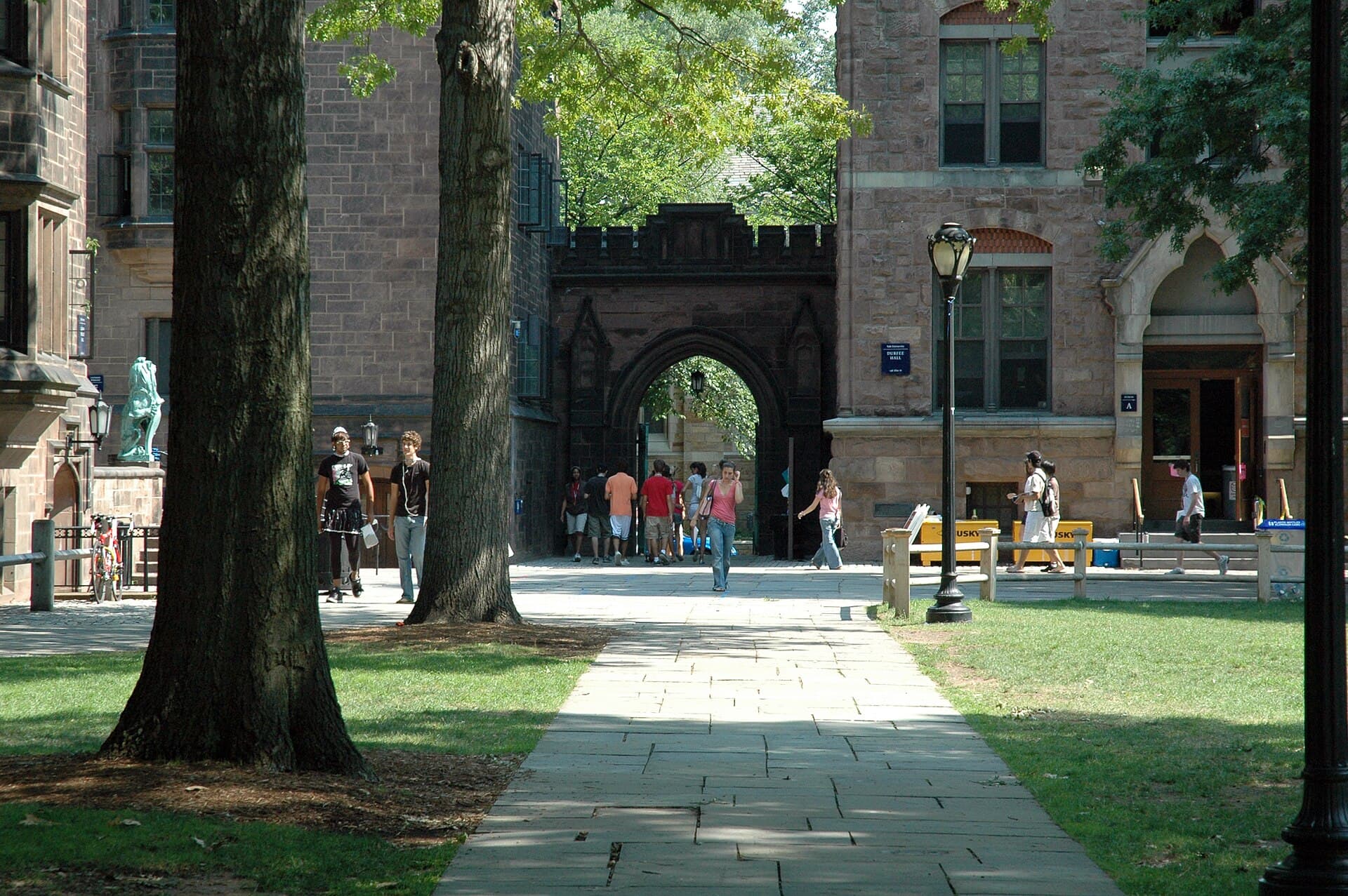 A tree-shaded path across Yale's Old Campus toward brownstone Durfee Hall and an arched gateway, with students walking