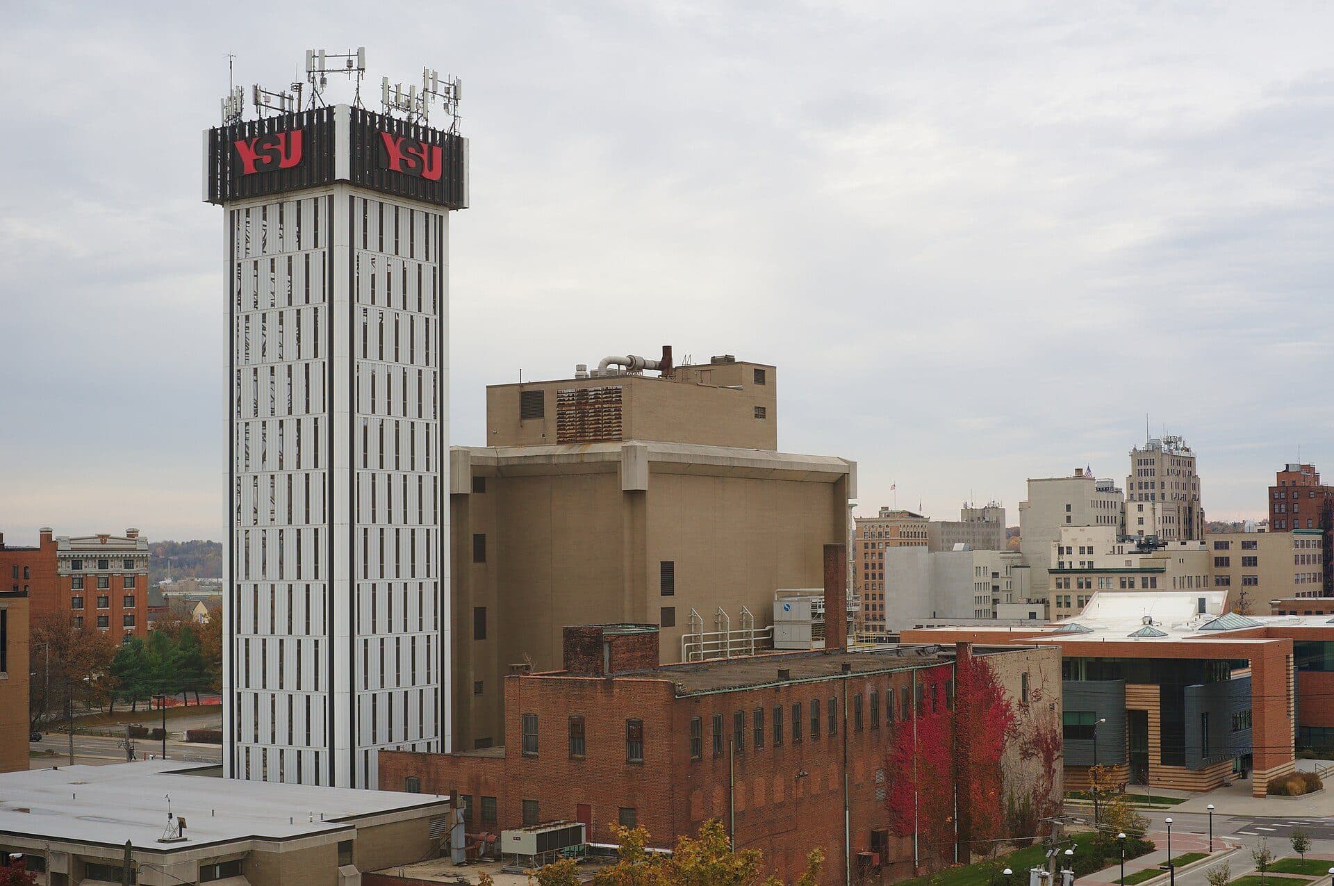 YSU's tall white clock tower with red YSU lettering rising above brick buildings and downtown Youngstown under a cloudy sky