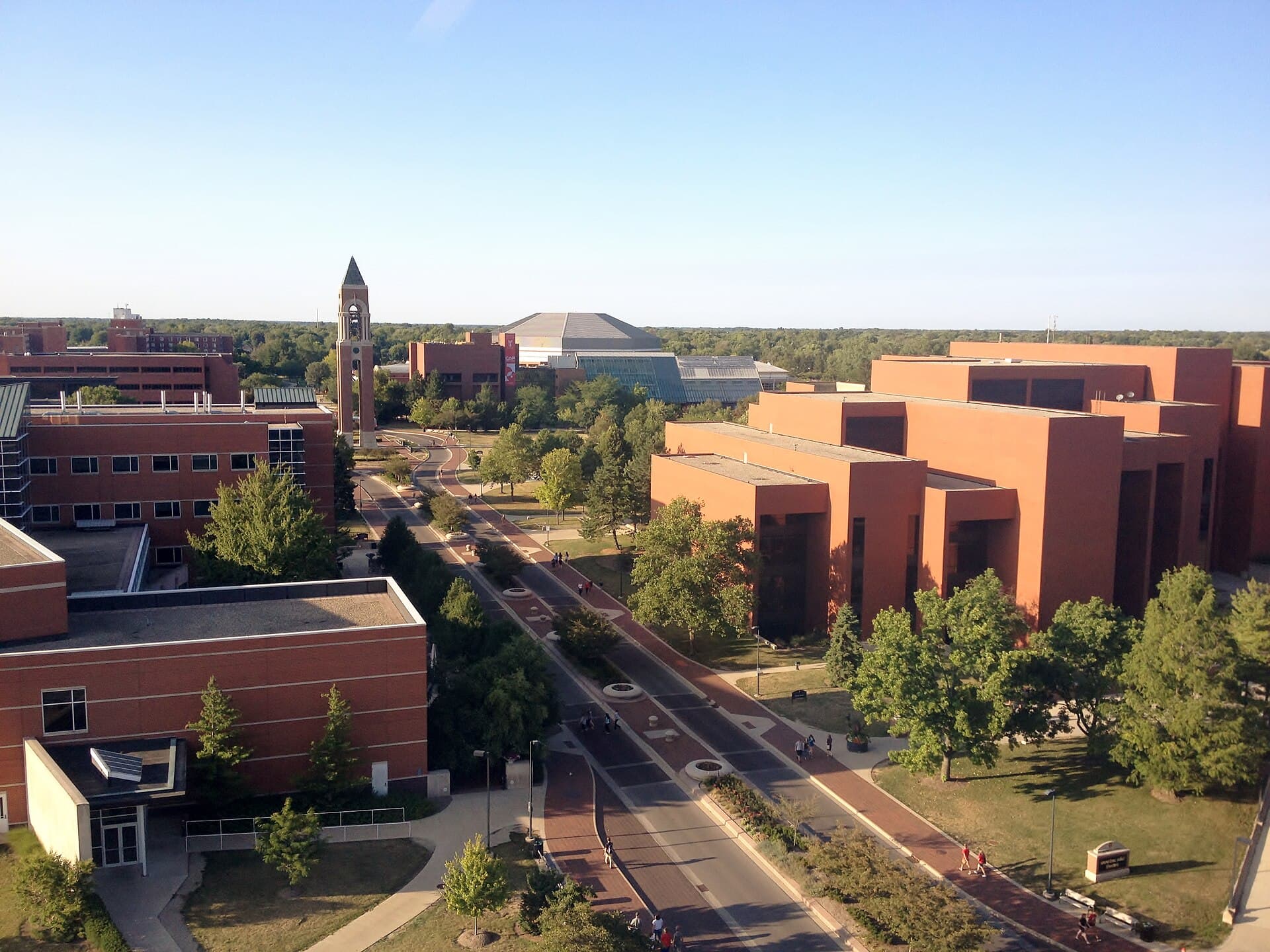 Looking down on Ball State's campus on a clear summer evening: a walkway-lined road curves between red-brick buildings and green trees toward a tall brick bell tower, with a large domed arena and flat Indiana farmland on the horizon