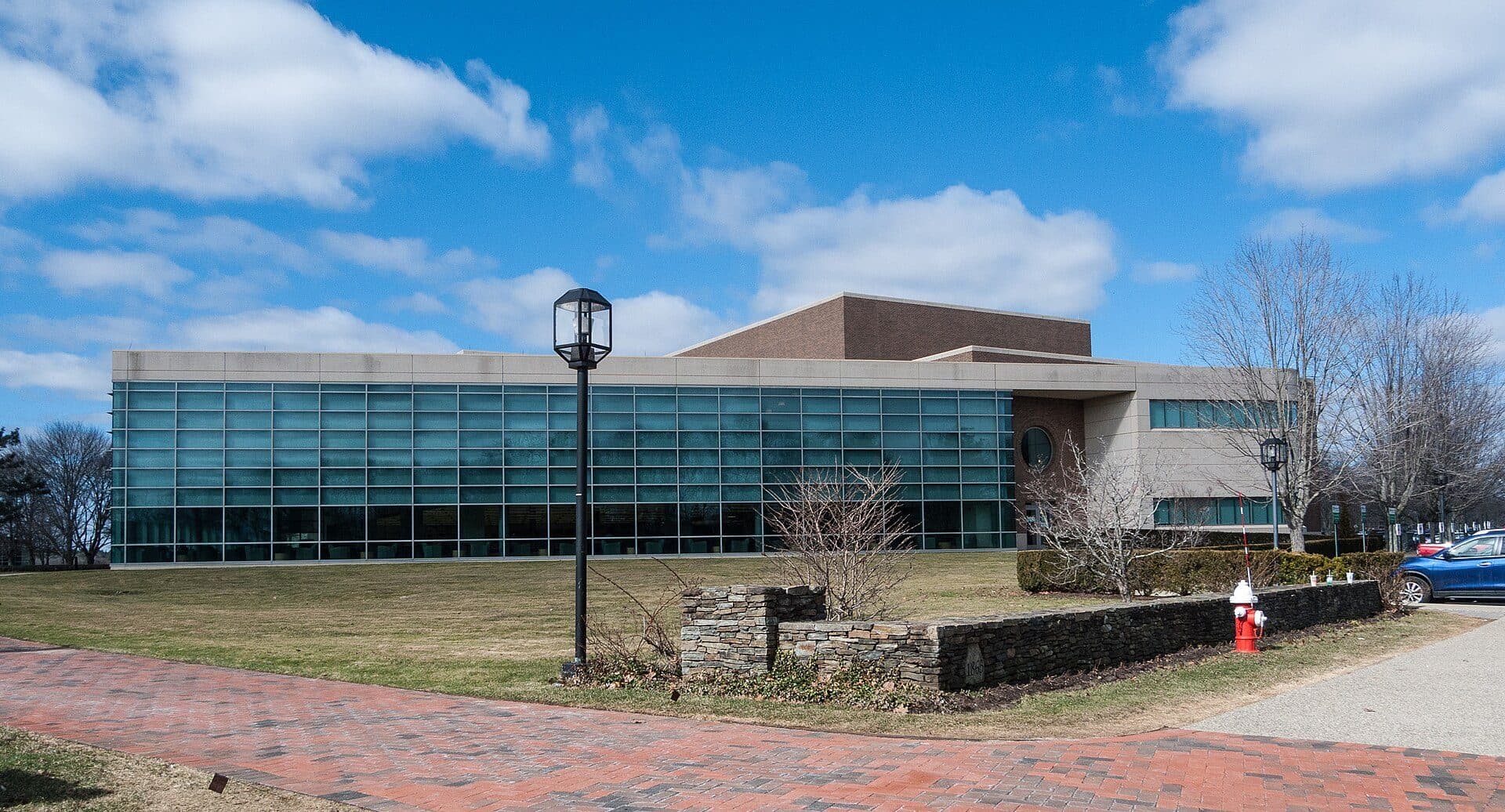 Bryant University's Bello Center, a long modern building with a curving blue-glass facade, seen across a brick walkway and a lawn on a sunny early-spring day