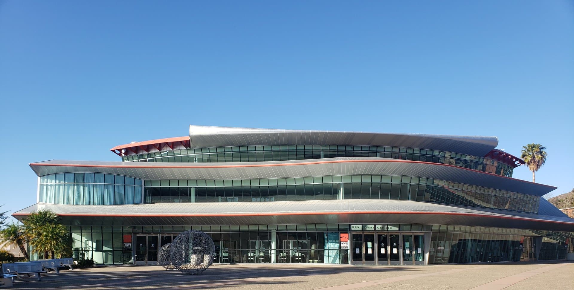 The curving glass and silver-metal Performing Arts Center at Cal Poly in San Luis Obispo, with red trim, palm trees and a round open-wire sculpture on the plaza under a clear blue sky