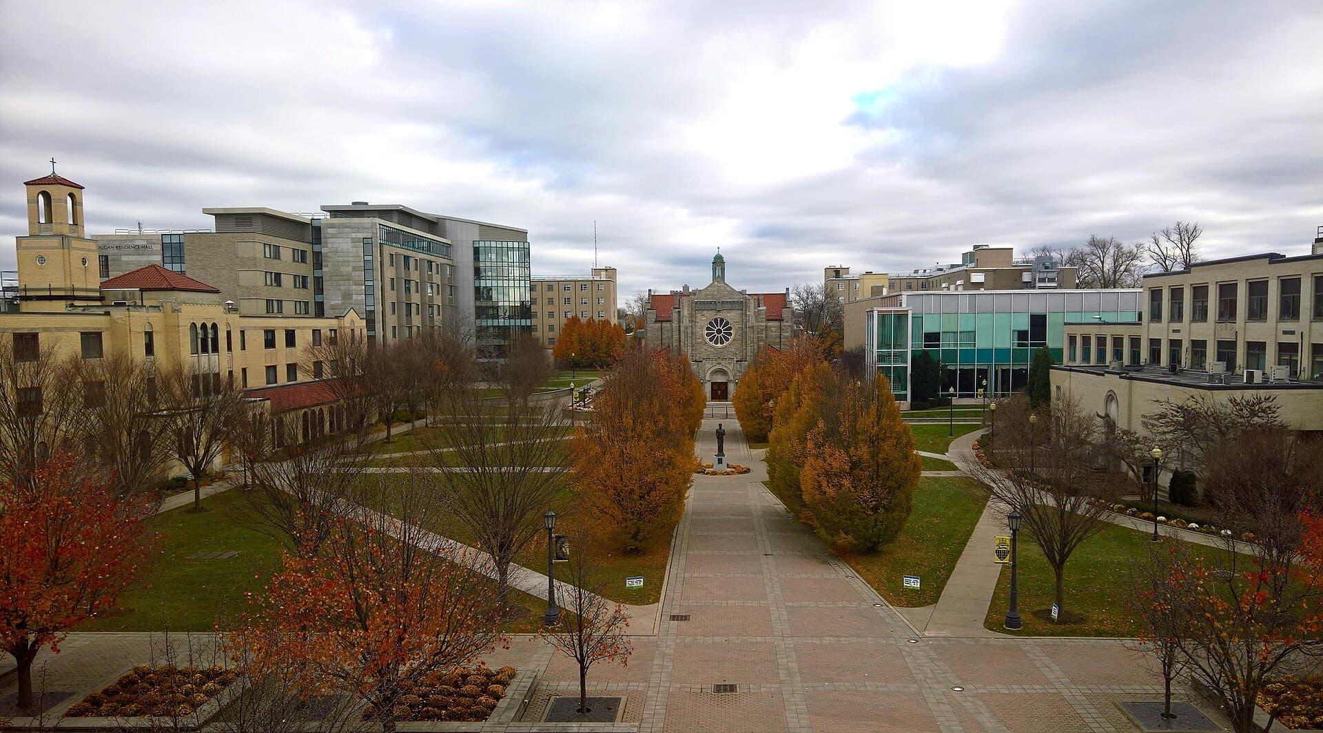 The Canisius quad seen from above in late fall, a brick-paved walk lined with orange and red trees leading to a stone chapel with a rose window, between tan and glass academic buildings under a gray sky