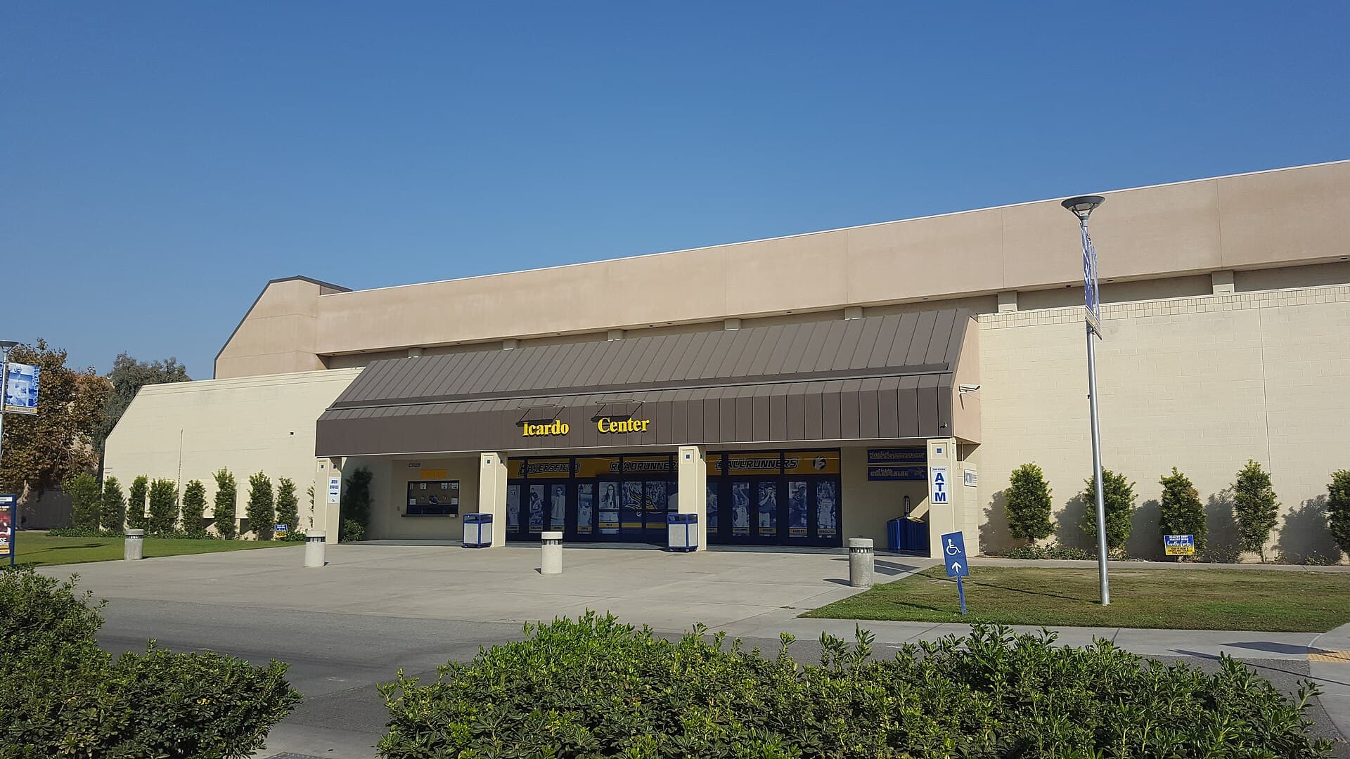 A long tan arena building with a brown metal awning over glass entrance doors, gold letters reading Icardo Center, trimmed shrubs and a lamppost in front under a clear blue sky