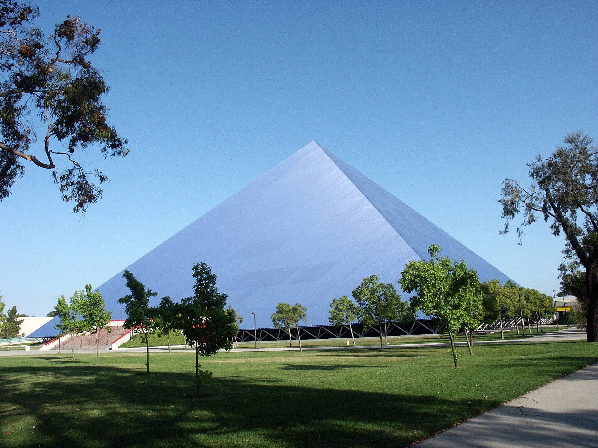 The blue Walter Pyramid arena at Cal State Long Beach rising behind a lawn and young trees under a clear sky