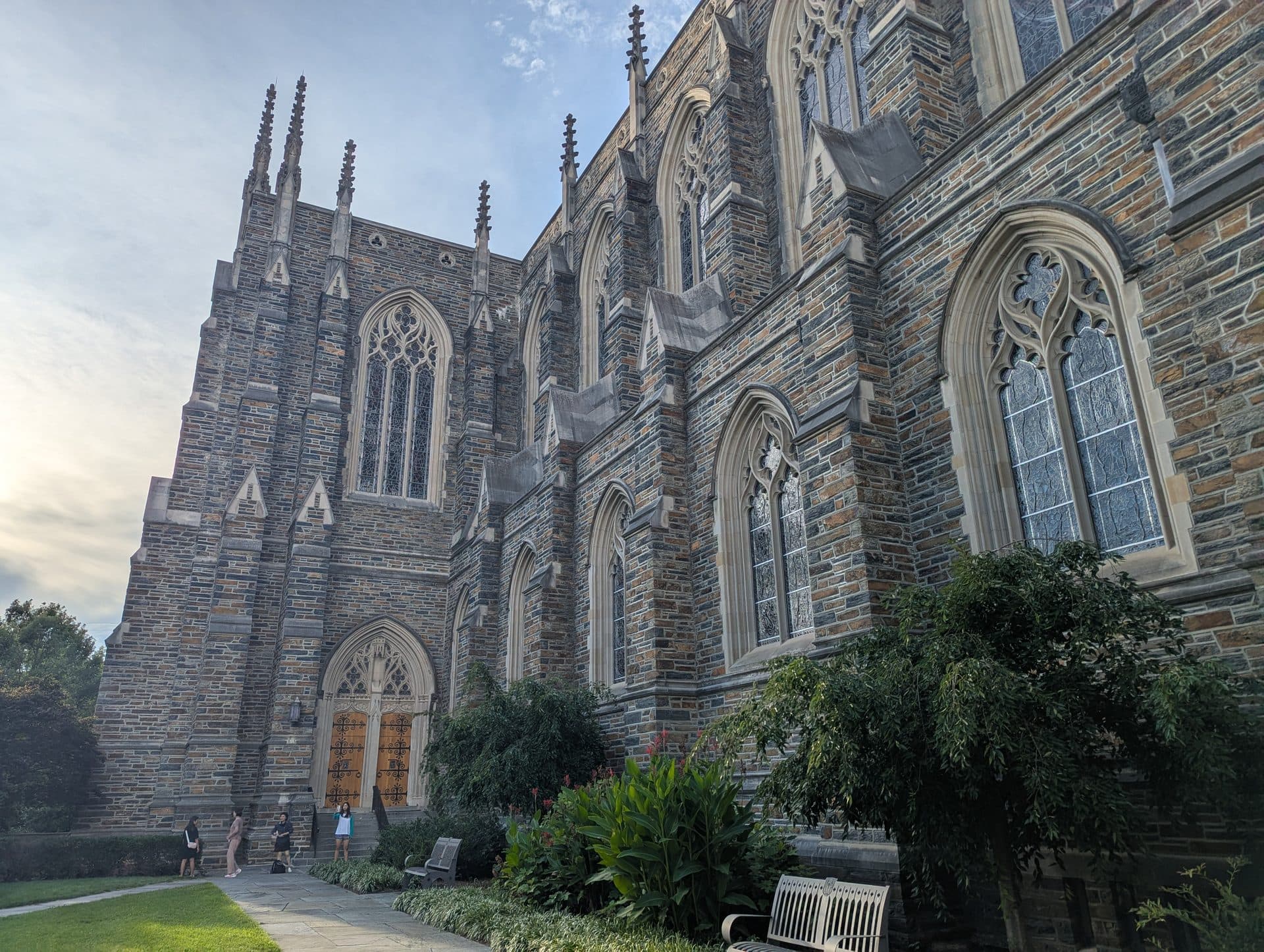 The side of Duke Chapel, a Gothic stone church with tall pointed stained-glass windows and spires, above a garden path with benches