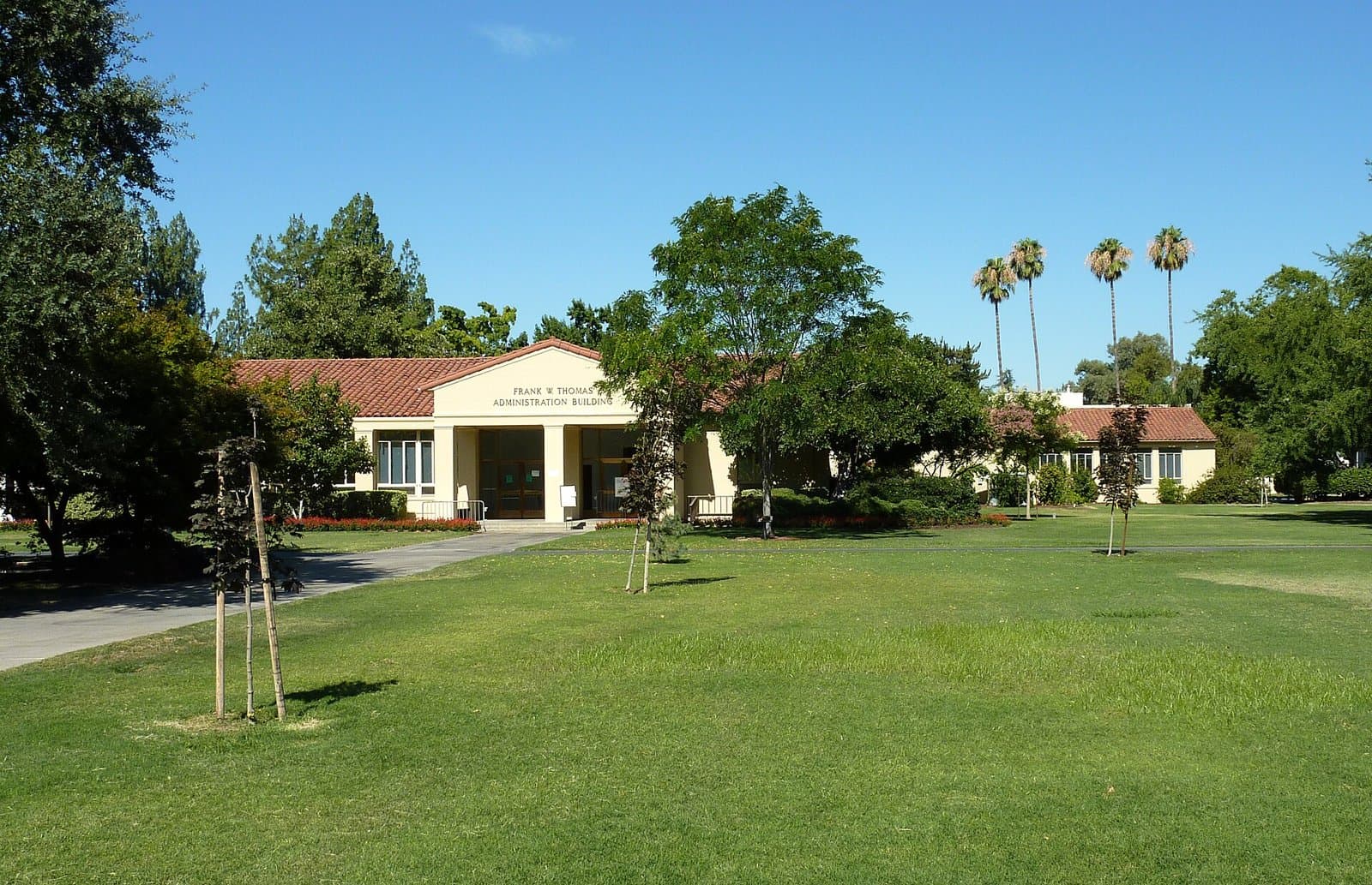 A single-story cream building with a red tile roof and the words Frank W. Thomas Administration Building over its entrance, behind a wide green lawn with young trees and tall palms under a clear blue sky