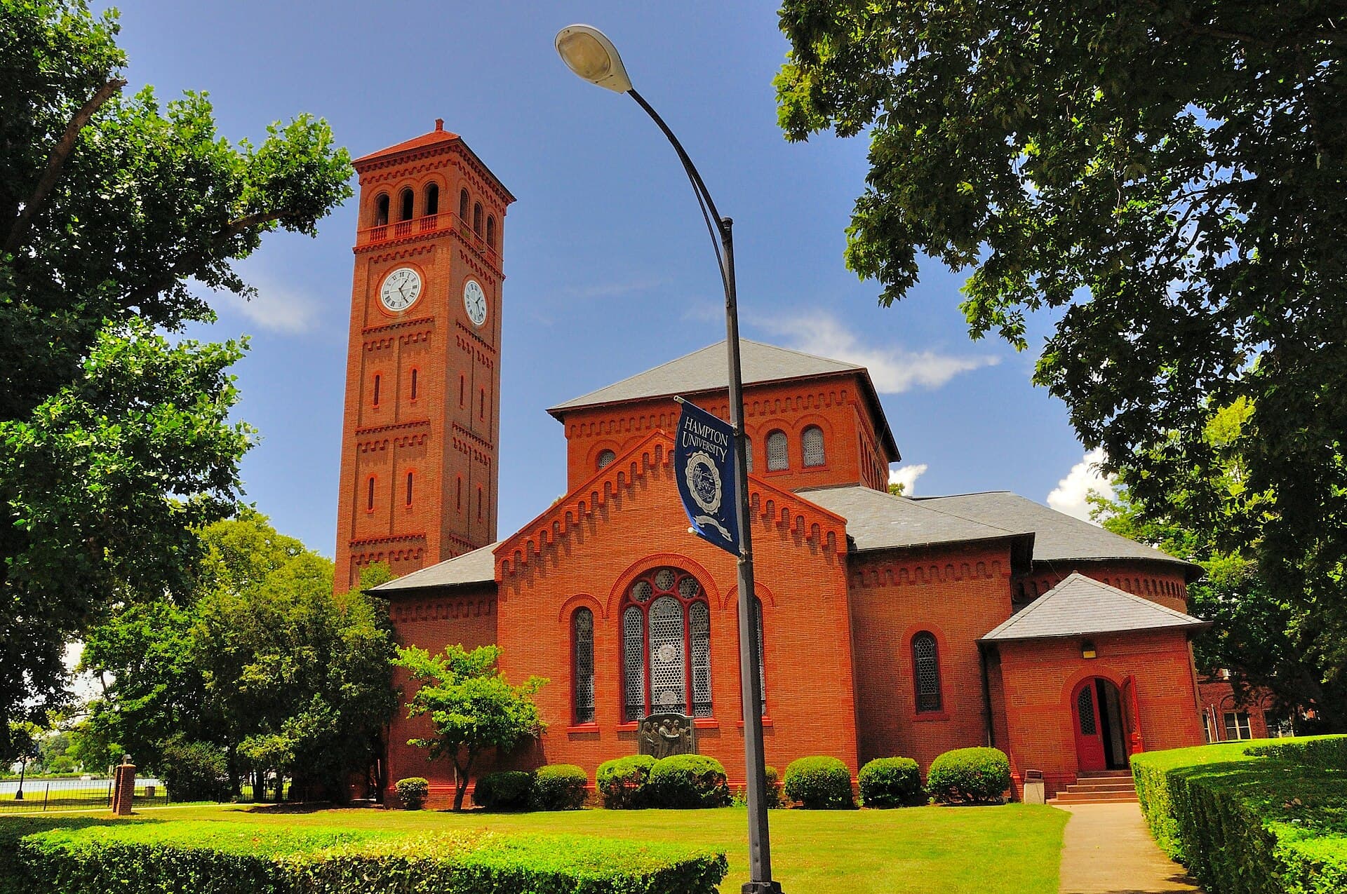 The red-brick Memorial Church with its clock tower at Hampton University, with a Hampton University banner on a lamppost, hedges and a lawn in front