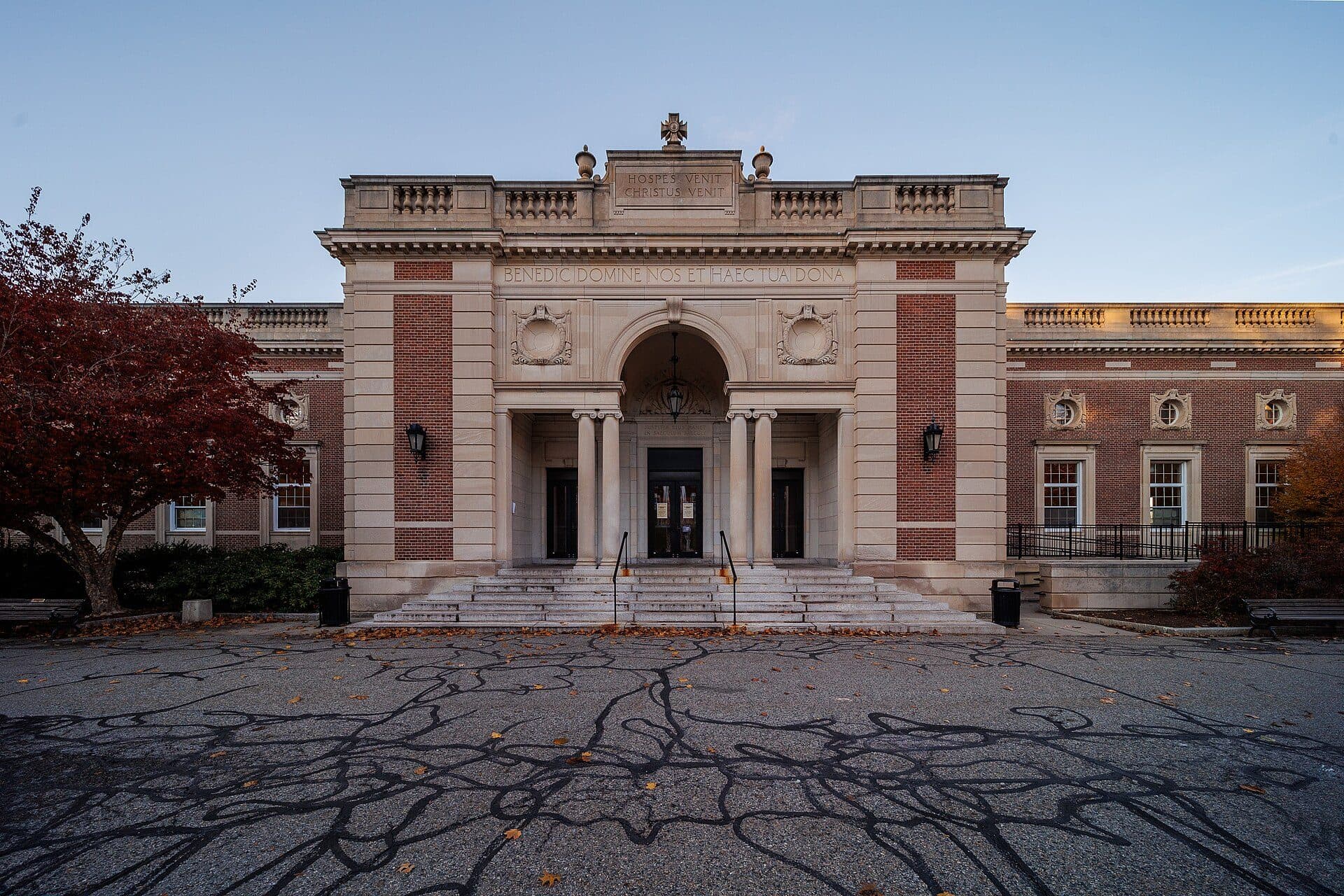 The brick-and-limestone front of Kimball Hall at Holy Cross, with a columned arched entrance, a carved crest above the doors and a red-leafed tree at left, behind a paved plaza