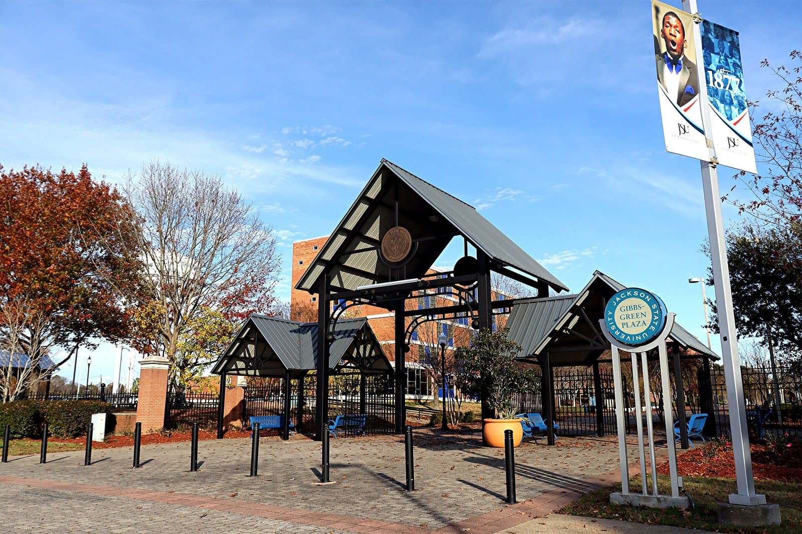 A paved plaza with a tall open timber-frame pavilion with a dark metal roof and a round medallion, two smaller pavilions with blue benches on either side, a round Gibbs-Green Plaza sign and a light pole with Jackson State banners, a red-brick building and autumn trees behind under a blue sky